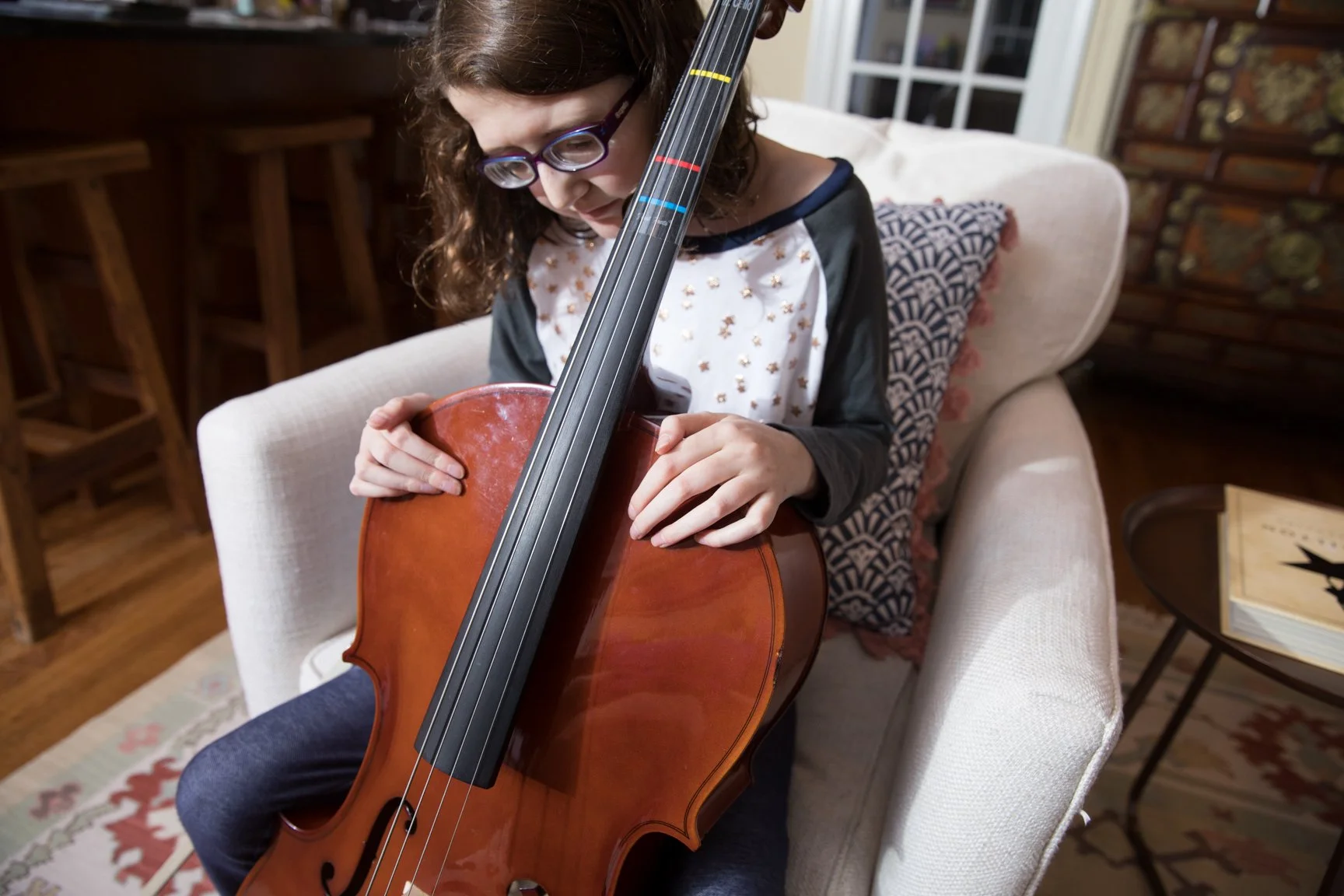 A young girl with glasses sitting on a beige armchair playing a violin, holding the bow and the violin close to her face.