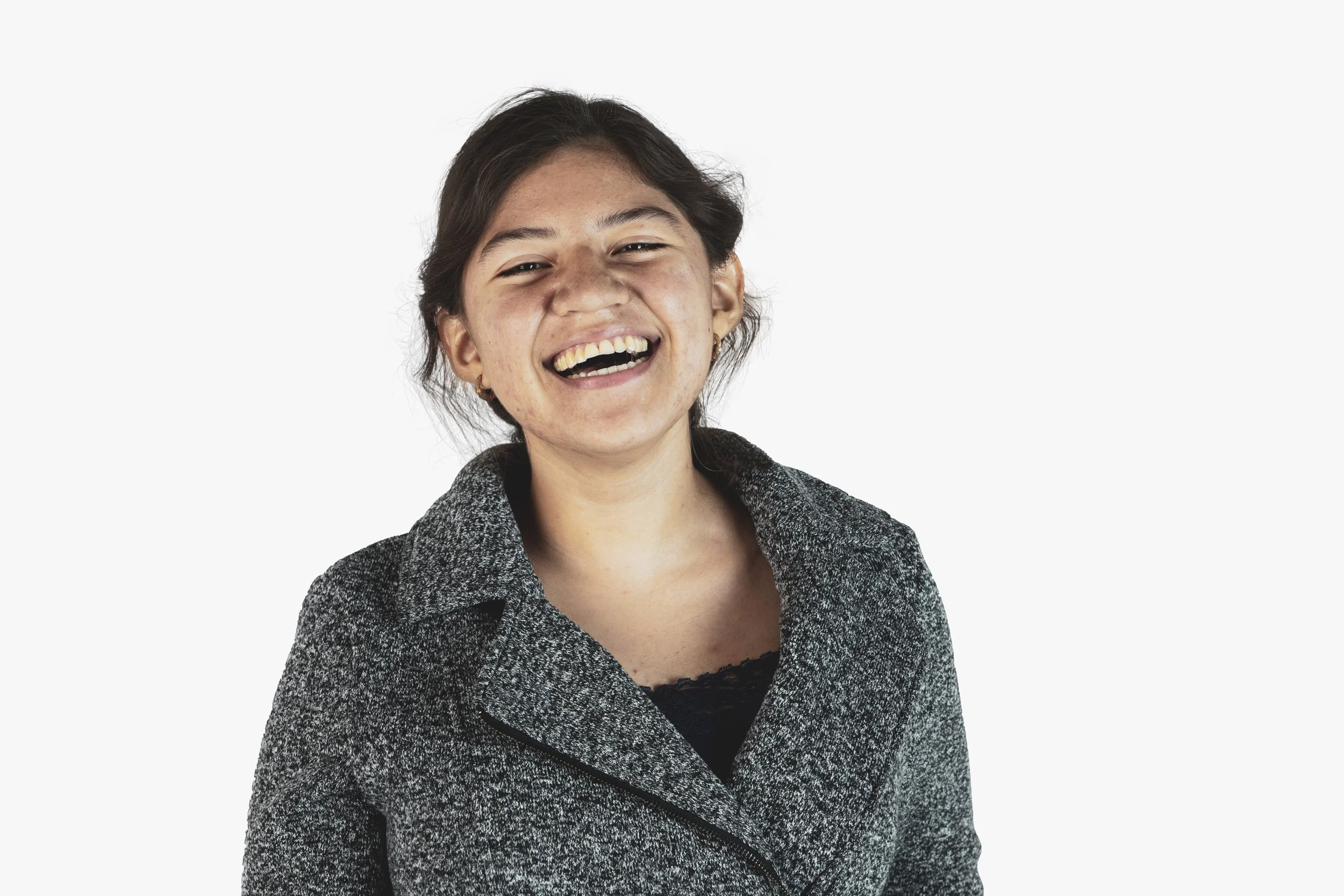 Young woman with dark hair smiling and laughing, wearing a gray textured blazer over a black top, against a plain white background.