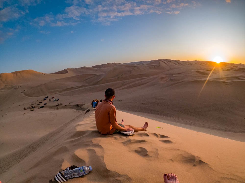 Person sitting on sand dune in desert at sunset, with other people in the background, footprints in the sand, and a water bottle with shoes nearby.