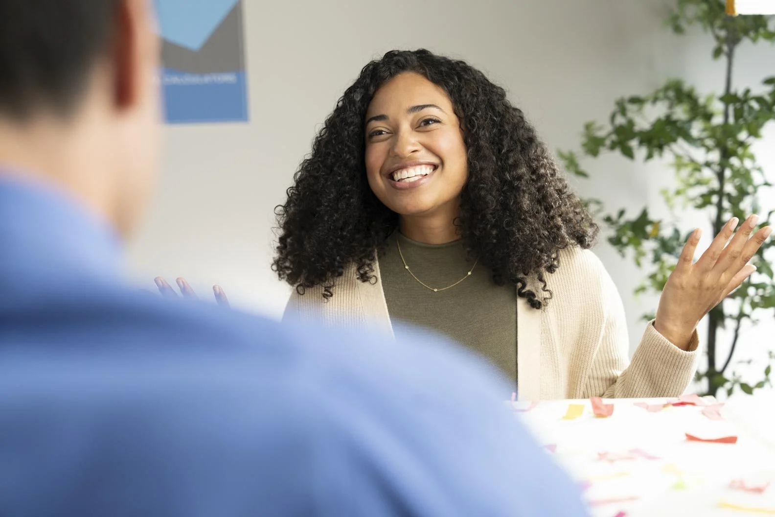 A smiling woman with curly hair talking to a person in blue, seated at a table with colorful notes or paper