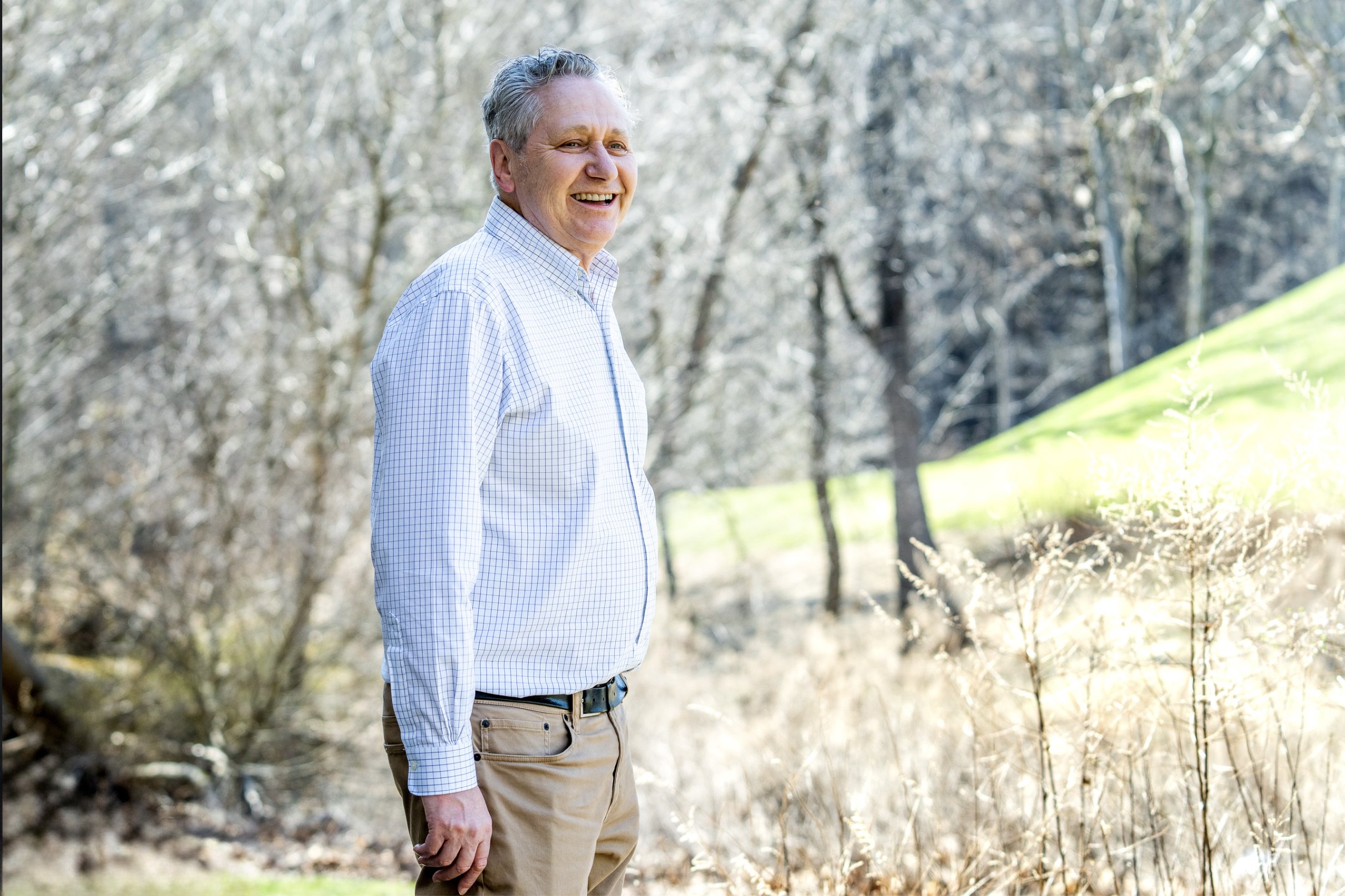 A smiling older man with gray hair standing outdoors in a sunlit, wooded area during late fall or early winter, wearing a light checkered shirt and khaki pants.