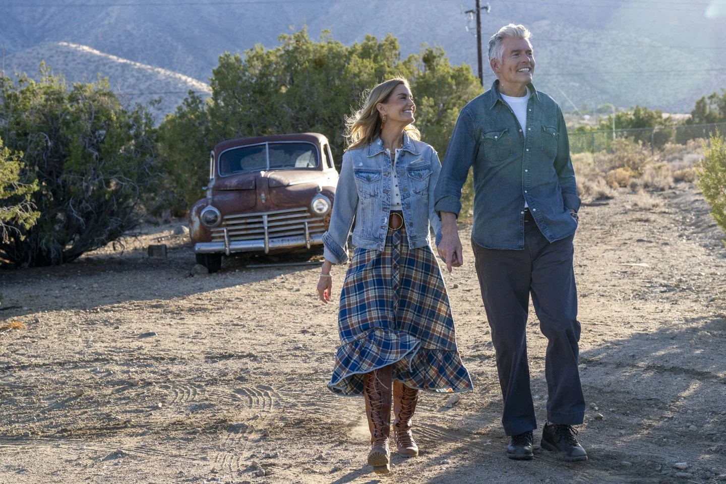 A man and woman walking in a desert area with a vintage car in the background, mountains, and clear sky.