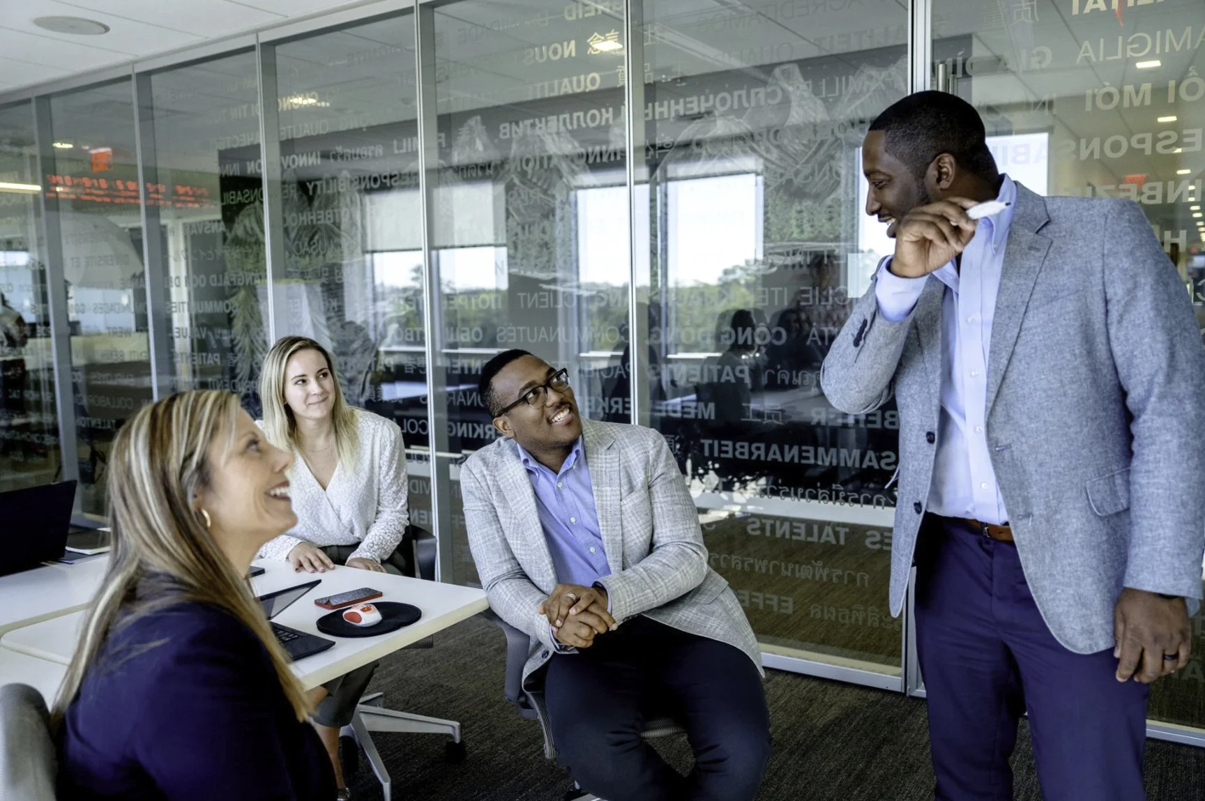 A diverse group of four professionals in a business meeting room, with one man standing and talking while three colleagues seated and listening attentively, all dressed in business attire.