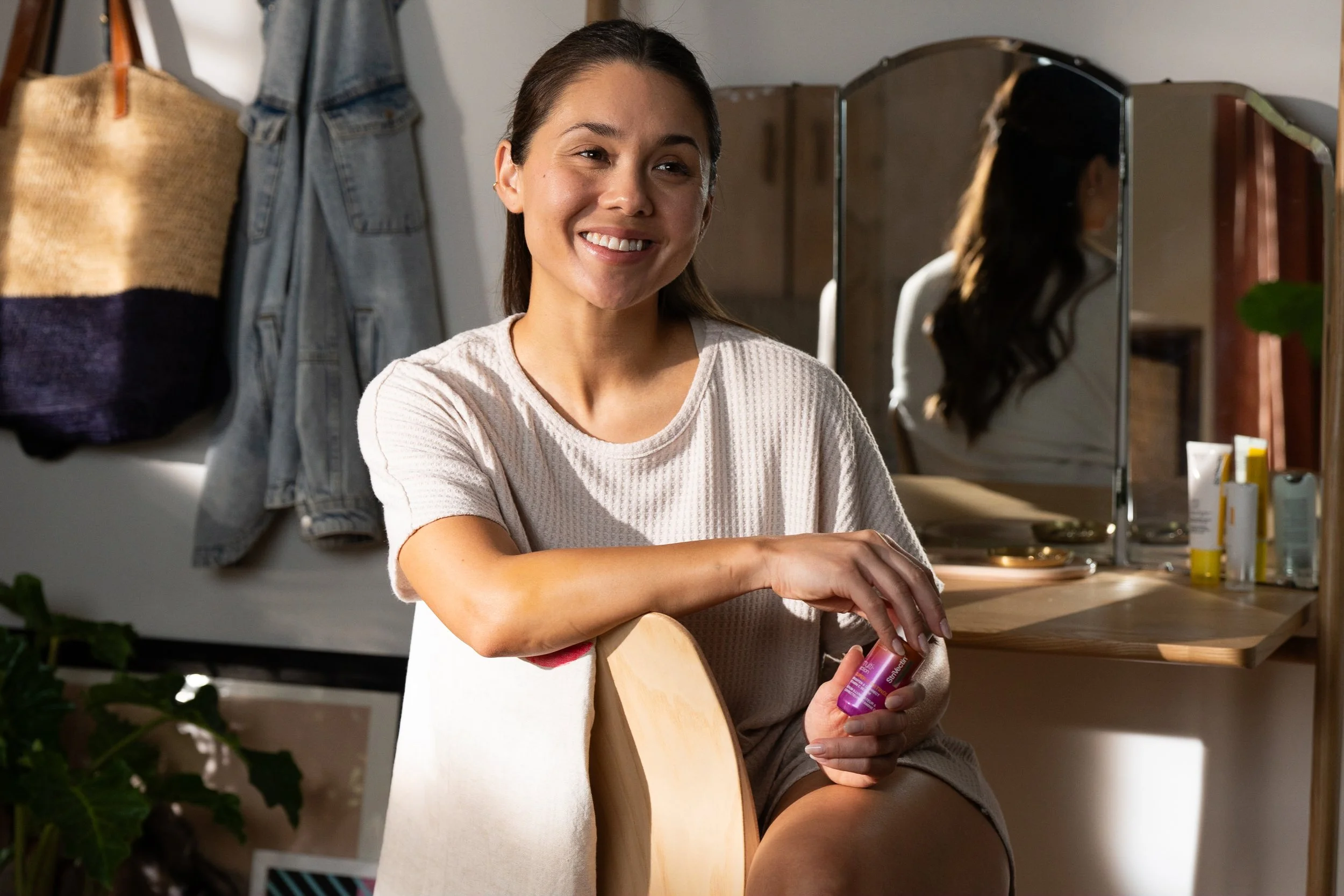 A woman sitting in front of a mirror, smiling, holding a small container of skincare product, in a room with a wooden vanity and various beauty items.