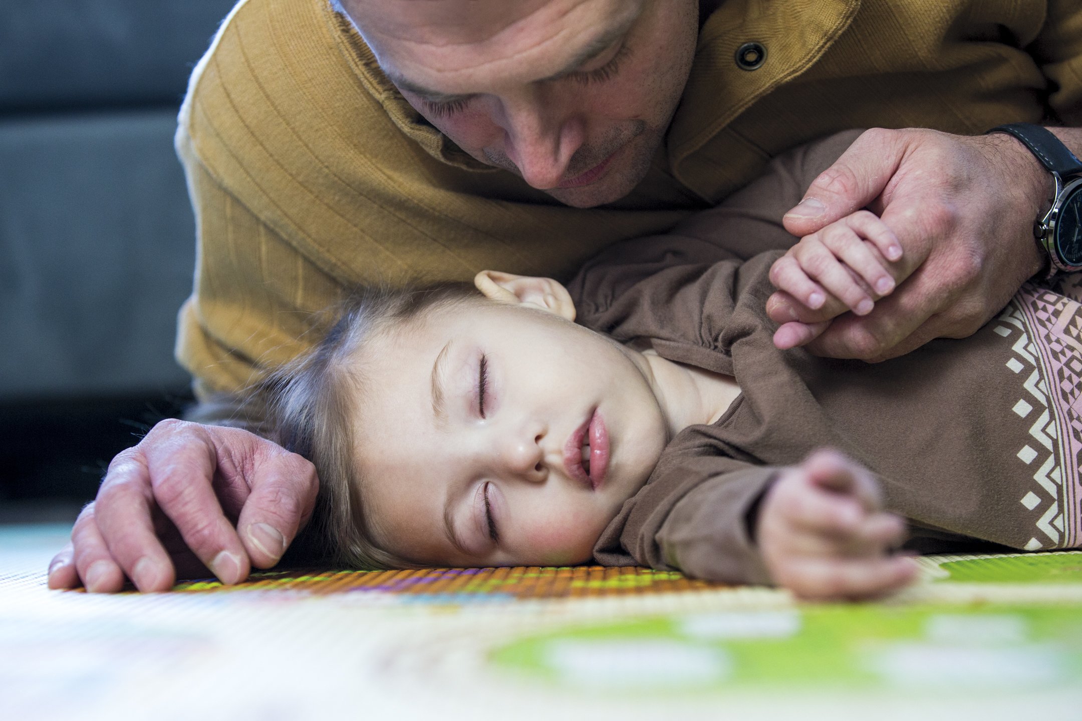 A man is lying on the floor with a young girl sleeping beside him, holding her hand gently. They are on a colorful mat and appear to be resting peacefully.