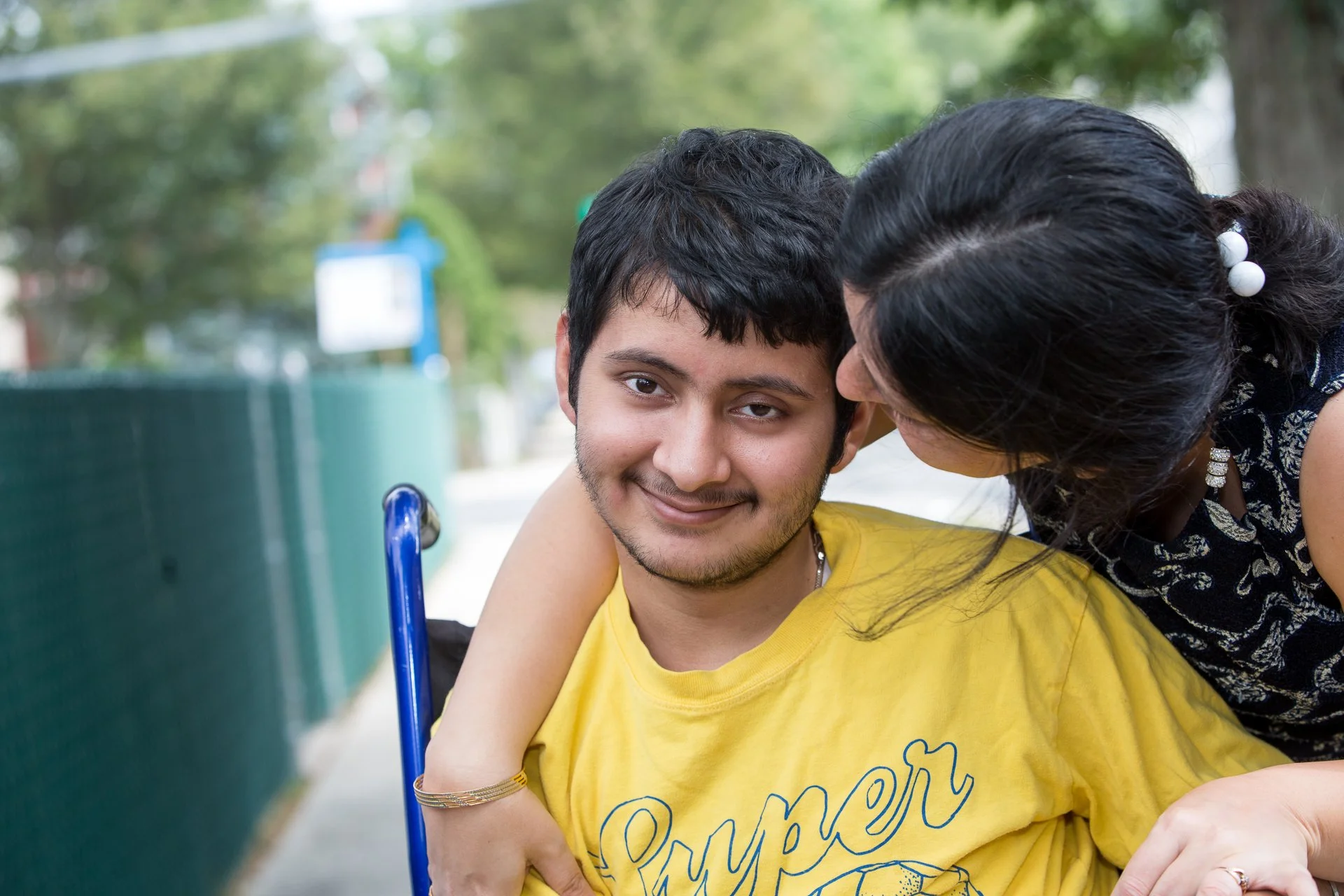 A young man in a yellow t-shirt sitting outdoors, being embraced and kissed on the temple by a woman with dark hair and white beads in her hair, on a sunny day with a green fence and trees in the background.
