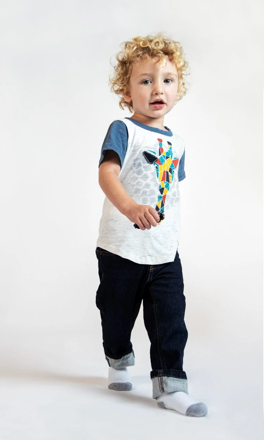 Young boy with curly blonde hair wearing a graphic giraffe t-shirt, dark rolled-up jeans, and white socks with gray toes, standing against a plain white background.
