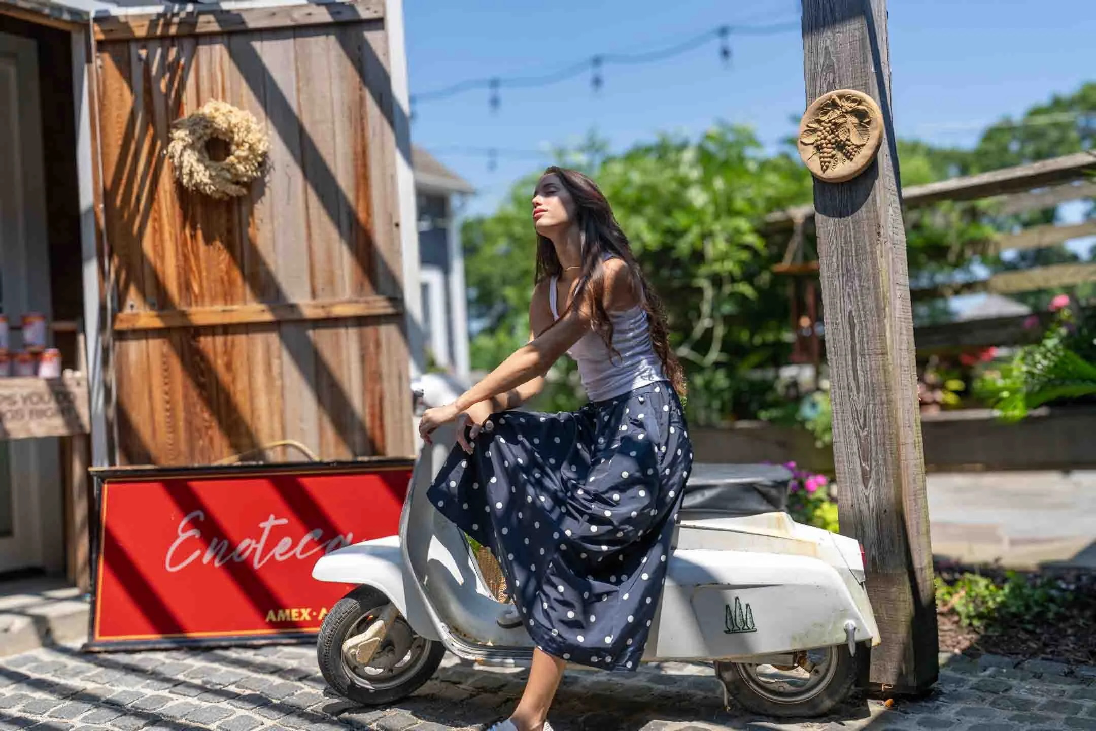 A woman in a white tank top and a navy blue polka dot skirt is sitting on a vintage white scooter parked on a cobblestone street. She is outdoors in a sunny setting with a wooden building, a red sign, and greenery in the background.