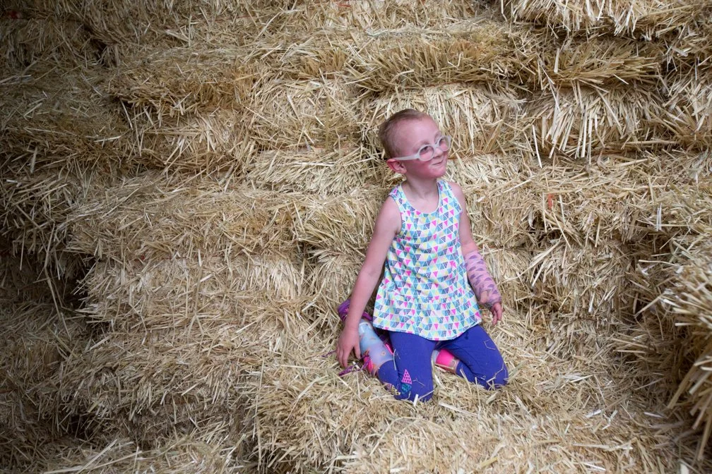 A young girl with glasses sits on hay bales in a barn or farm setting.
