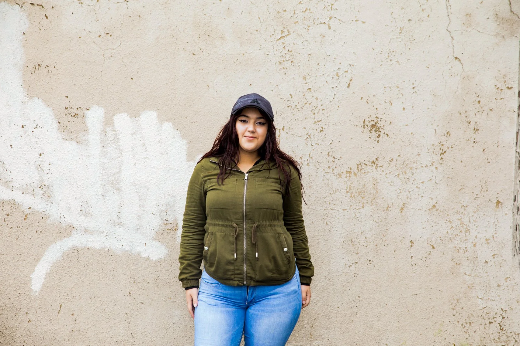 Young woman with dark hair wearing a black cap, an olive green jacket, and light blue jeans standing in front of a textured beige wall with some white paint splashes.