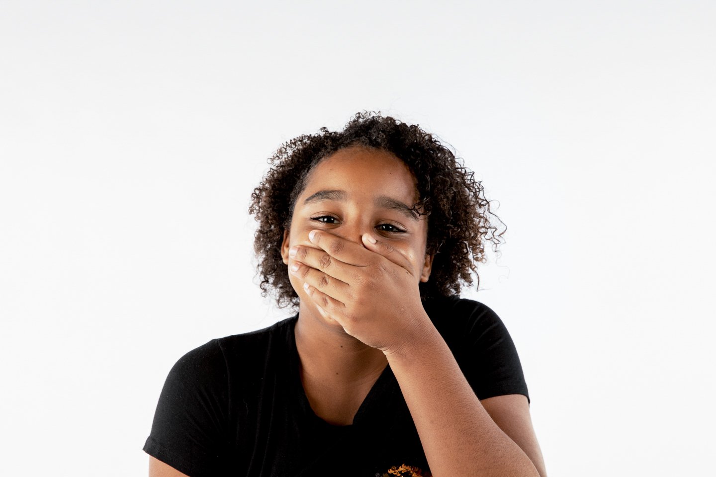 Young girl covering her mouth with her hand, smiling, against a white background.