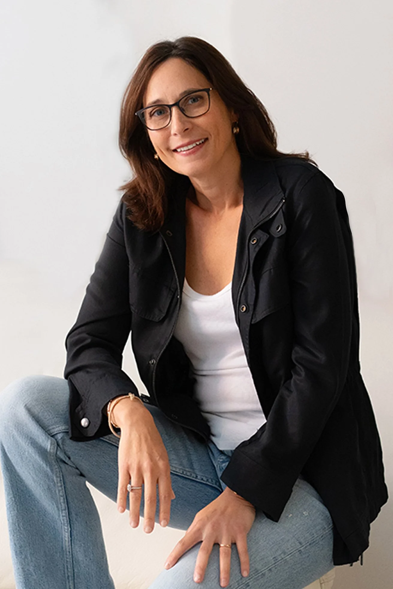A woman with brown hair, glasses, wearing a black leather jacket, white T-shirt, and light blue jeans, sitting against a plain white wall.