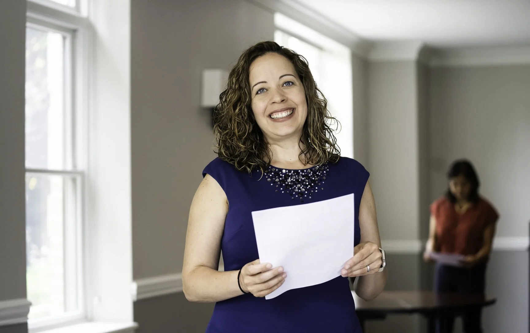 A smiling woman with curly brown hair wearing a navy blue dress with embellishments on the neckline holding a piece of paper, standing in a brightly lit room with gray walls and large windows.