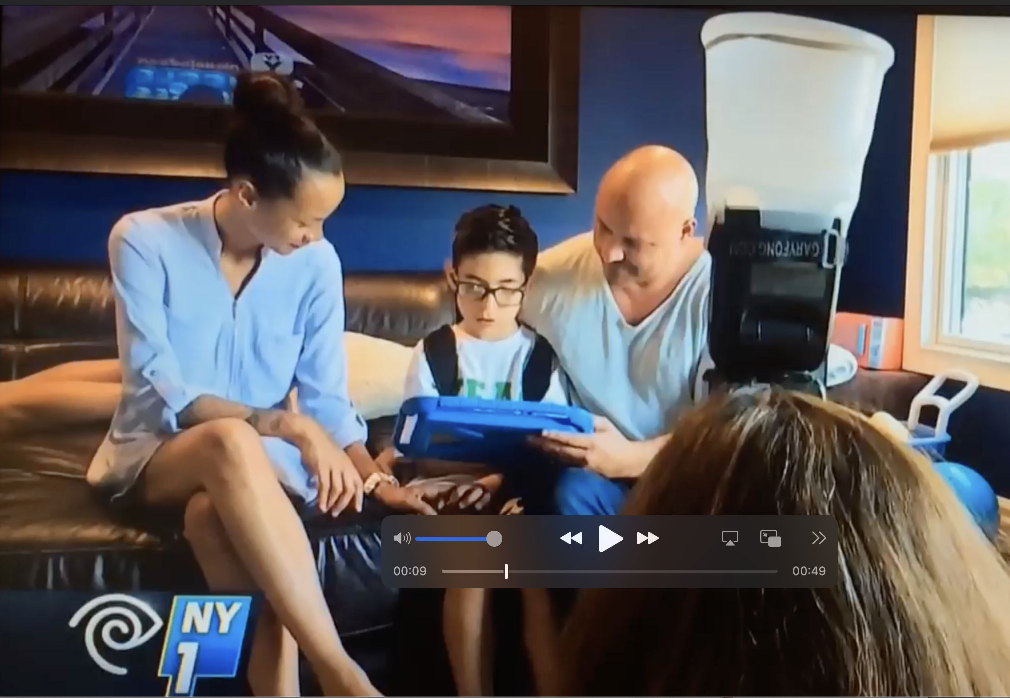 A girl in a light blue dress sitting on a black leather couch, smiling at a boy wearing glasses and a backpack, who is holding a blue electronic device, with a man in a white shirt sitting beside them, looking at the device. A woman with long brown h