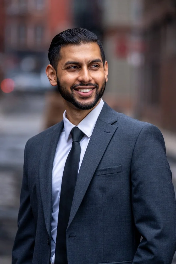 A smiling man in a suit and tie standing outdoors in an urban setting.