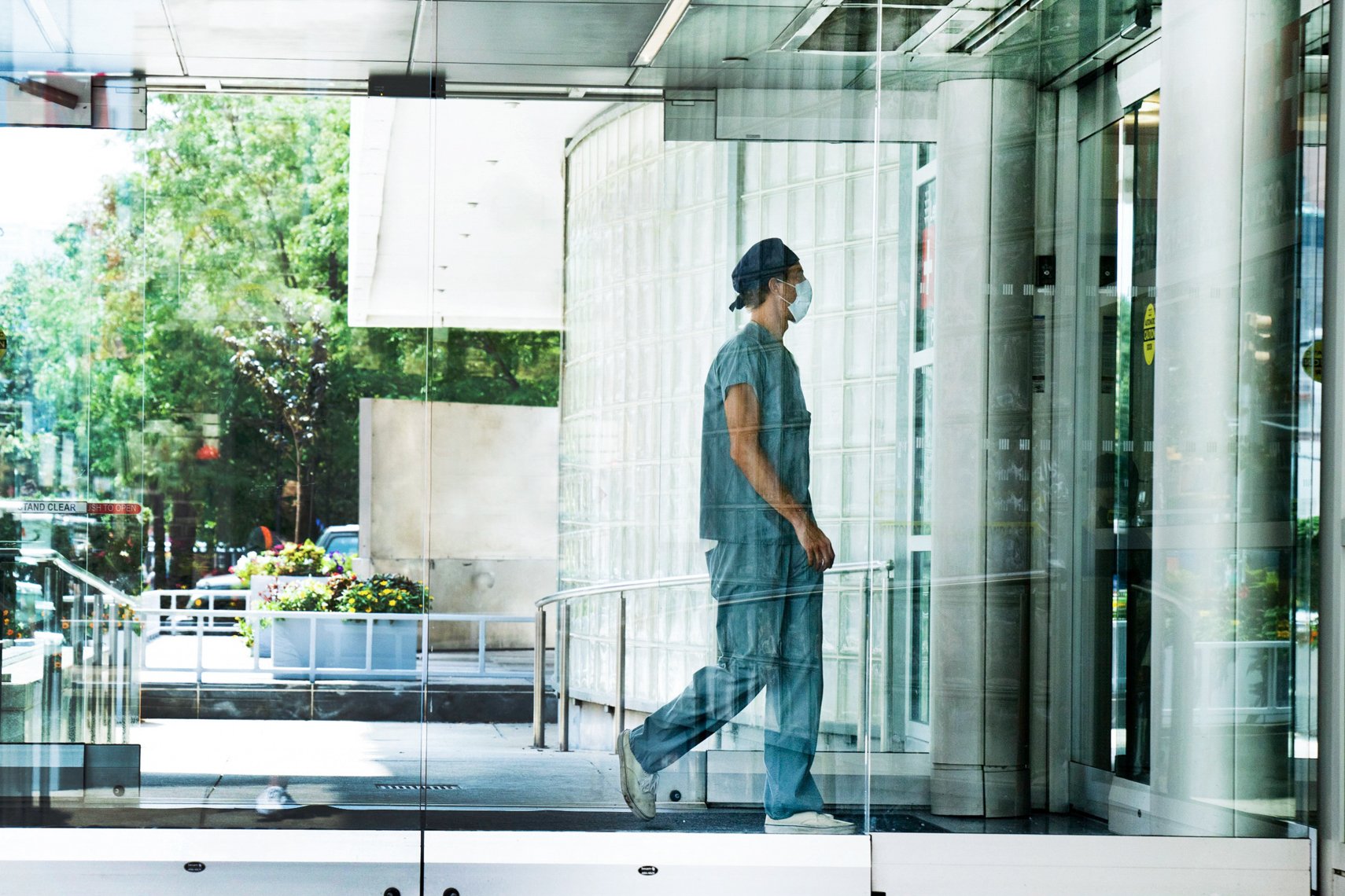 A person wearing medical scrubs, a face mask, and a hair cover, standing inside near glass doors, with trees and outdoor scenery visible through the glass.