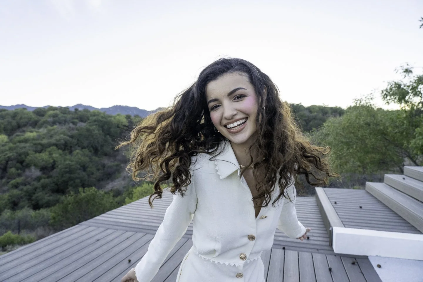 A young woman with long, curly hair smiling on a rooftop with mountains and trees in the background.