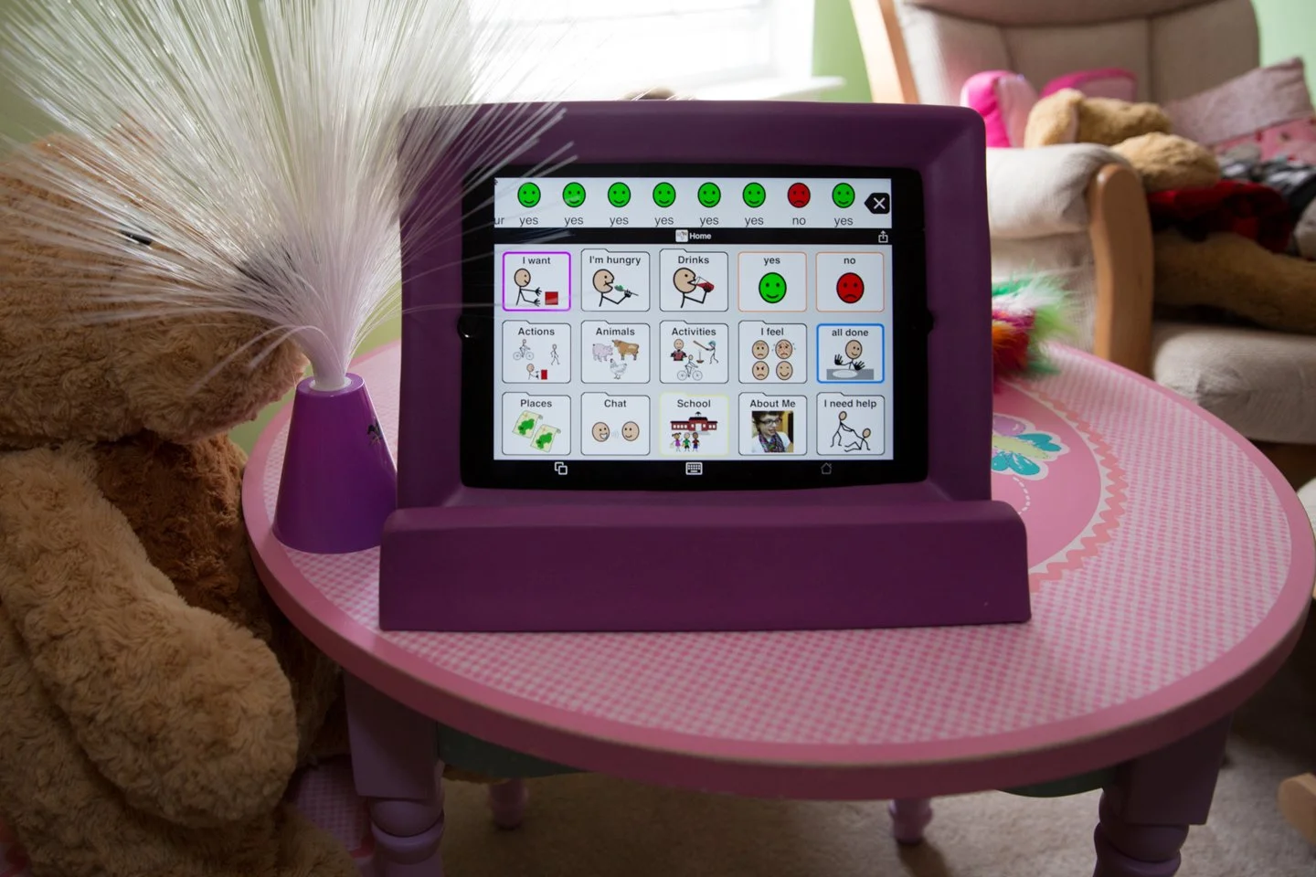 A tablet on a pink children's table displaying a communication app with various icons and options for assistance. The table has a teddy bear with a feather duster next to it.