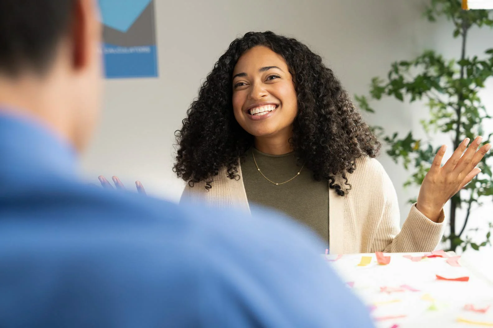 A young woman with curly hair smiling and gesturing during a conversation, sitting at a table with colorful confetti, in a well-lit indoor setting with a potted plant in the background.