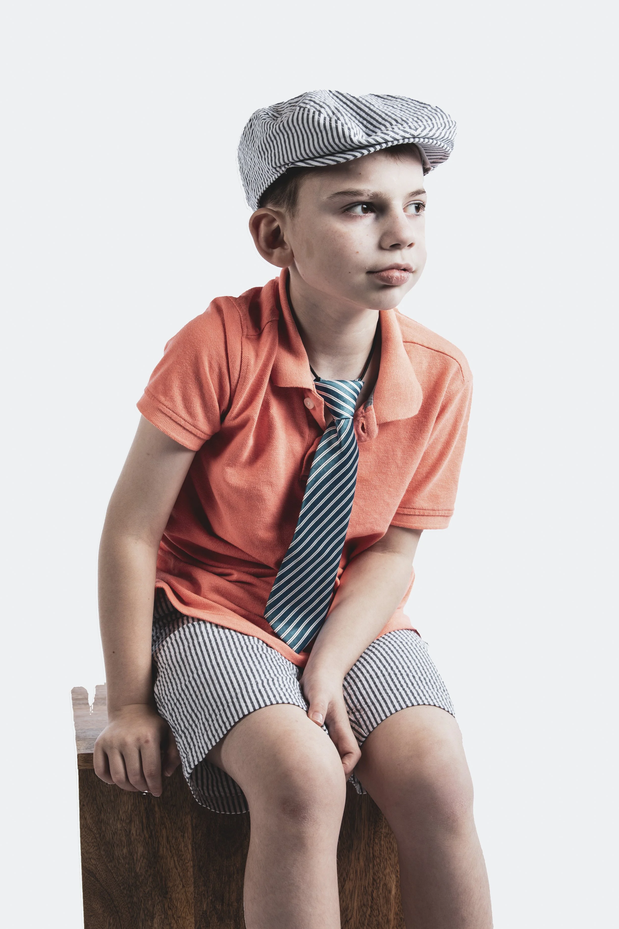 A young boy sitting on a wooden box, dressed in a casual outfit with a striped cap, coral T-shirt, striped shorts, and a striped tie, posing against a plain white background.
