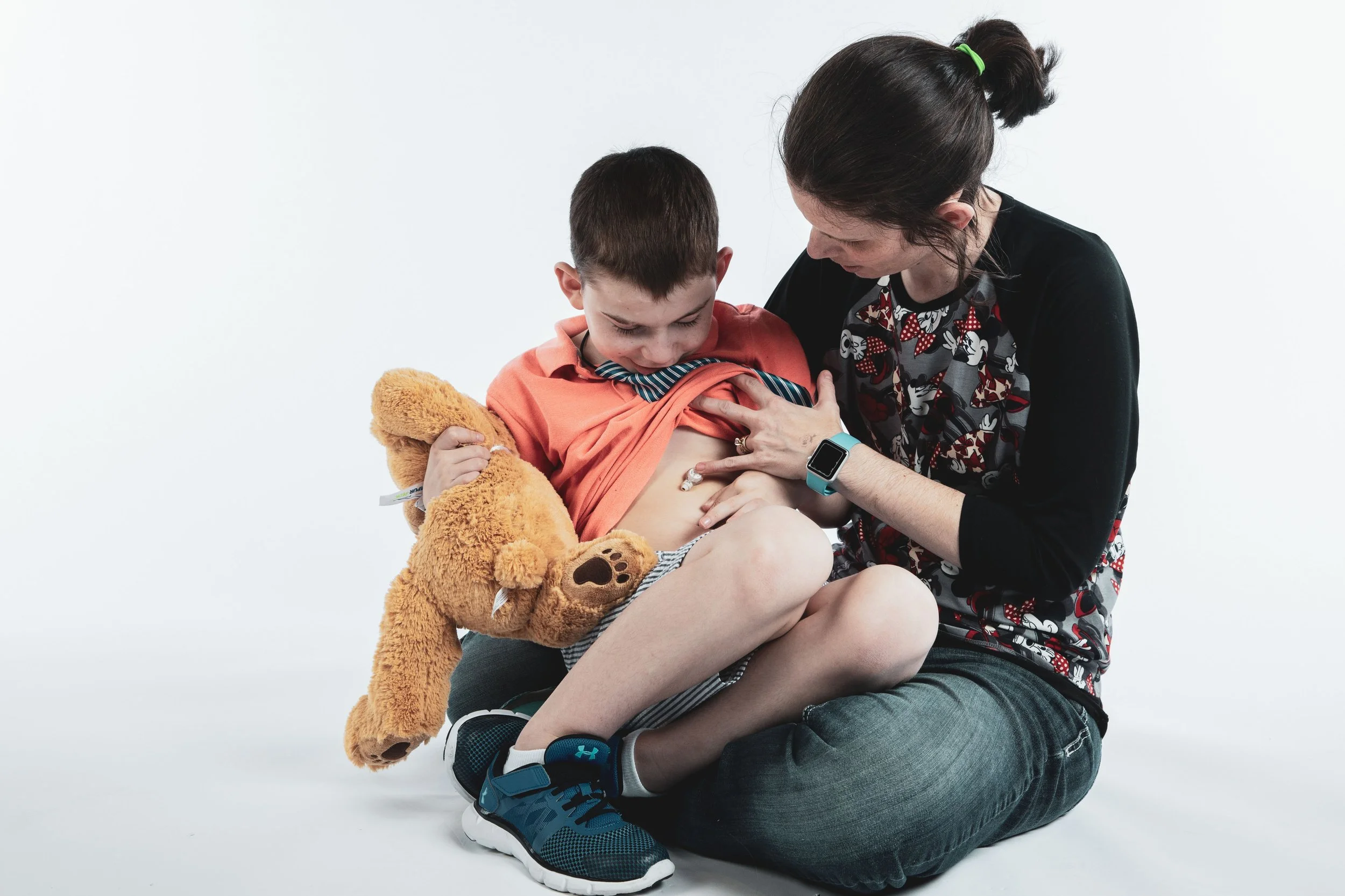 A woman helping a young boy look at his stomach, which appears to have a medical device or wound. The boy is holding a stuffed teddy bear and wearing sneakers, while the woman is close to him, wearing a patterned shirt and a smartwatch.