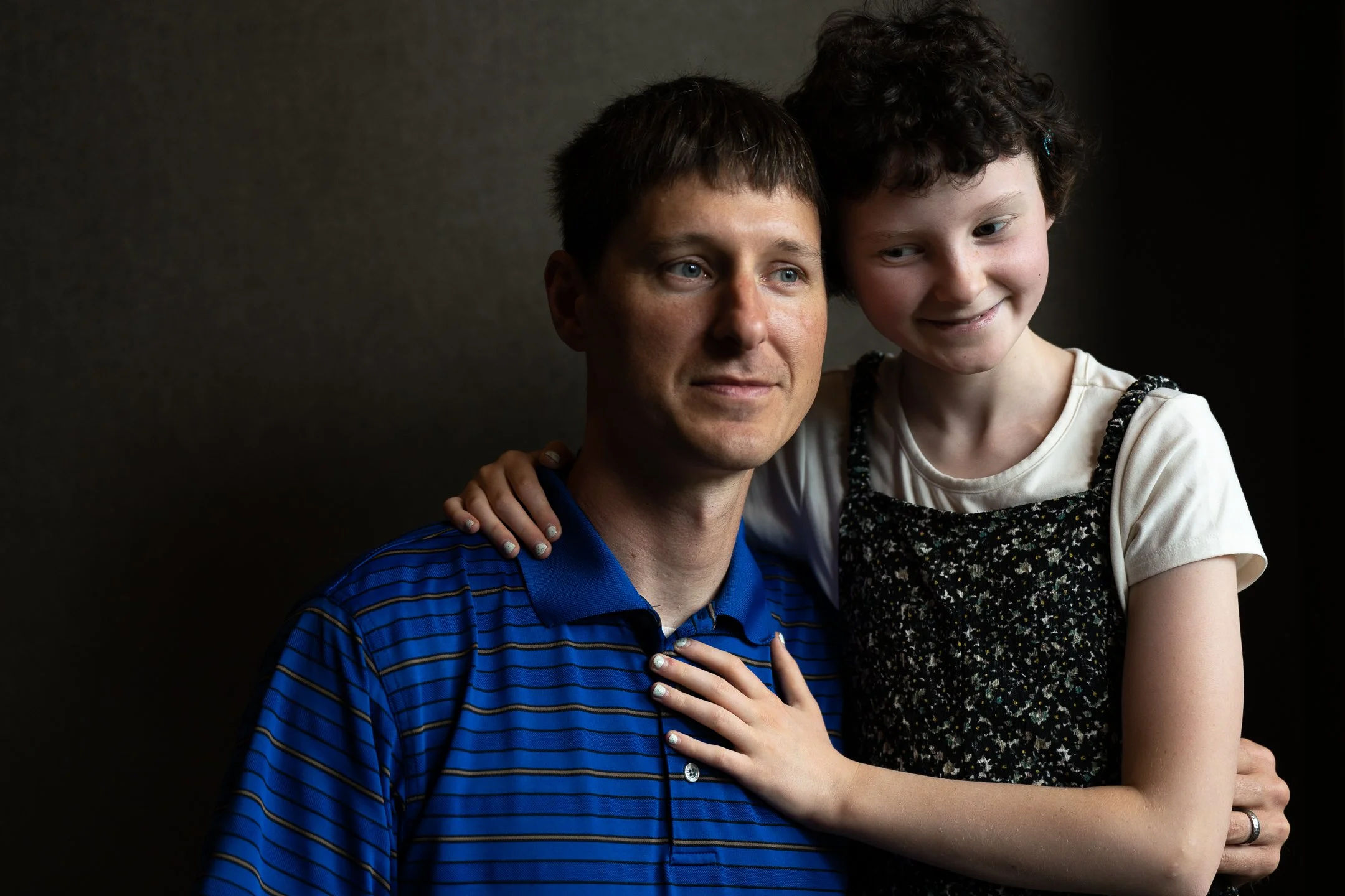 A man and young girl, likely father and daughter, pose together against a dark background. The girl has her arm around the man's shoulders and is smiling, while the man has a calm expression.