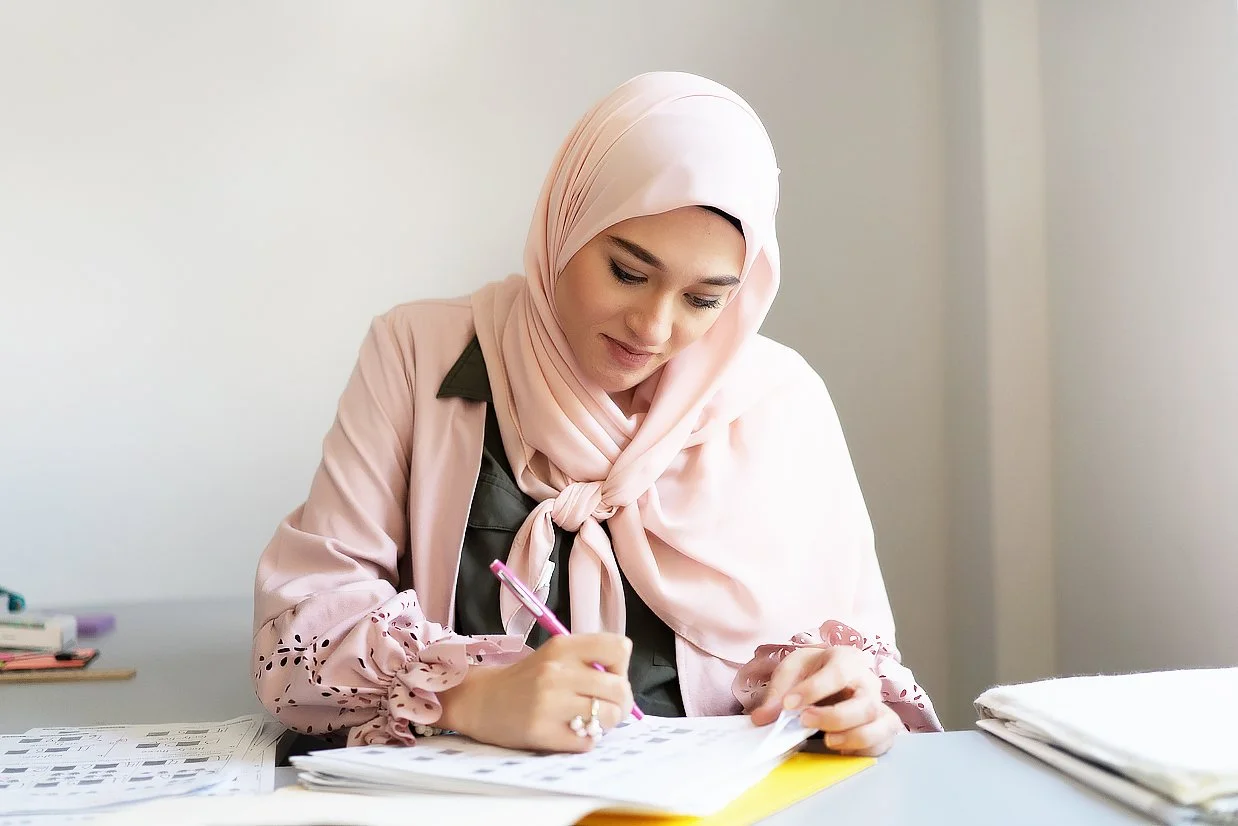 A woman wearing a light pink hijab and beige jacket sitting at a desk, writing in a notebook with a pink pen, surrounded by papers and folders.