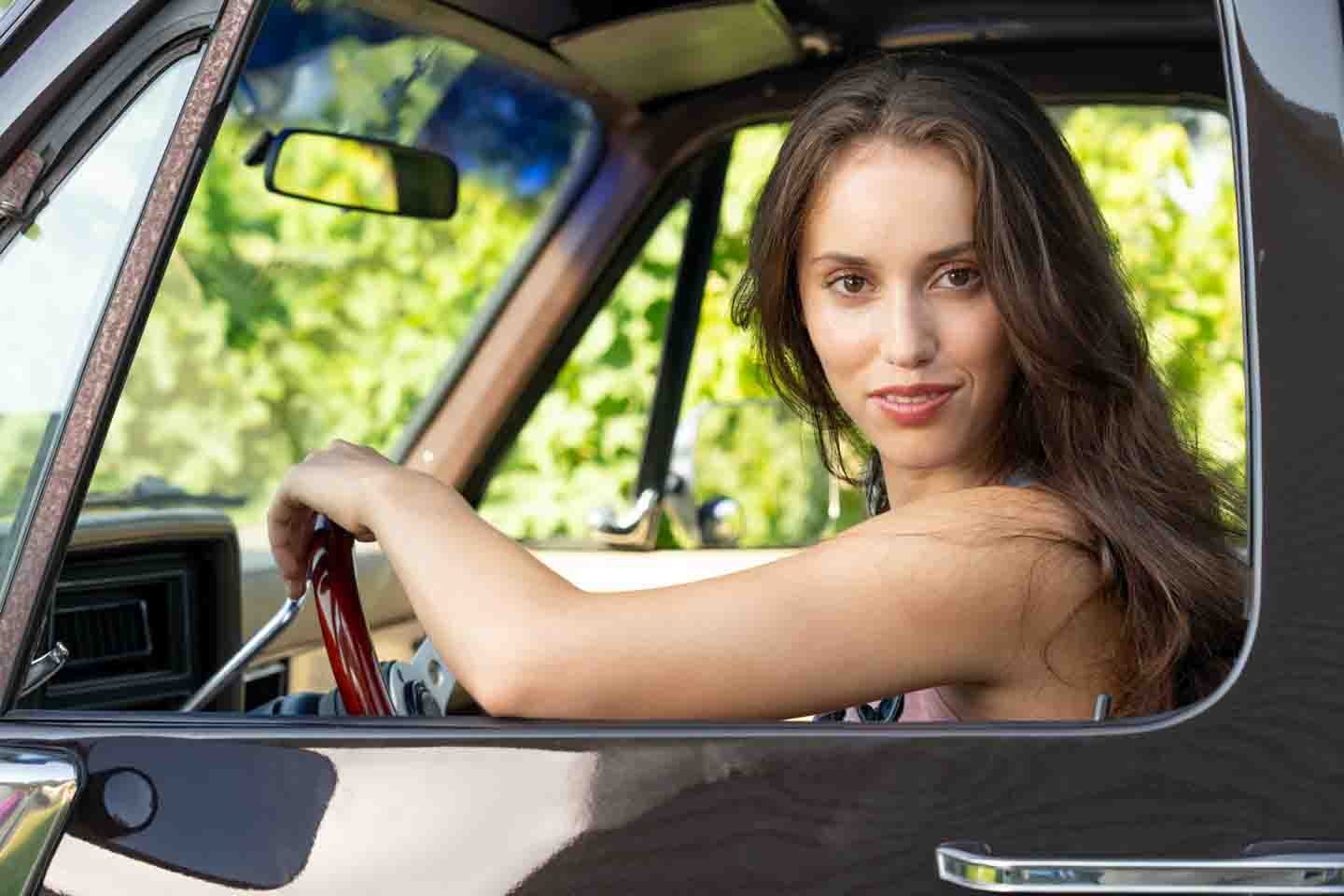 A young woman with long brown hair sitting in the driver's seat of a vintage car, looking out the window with a slight smile.