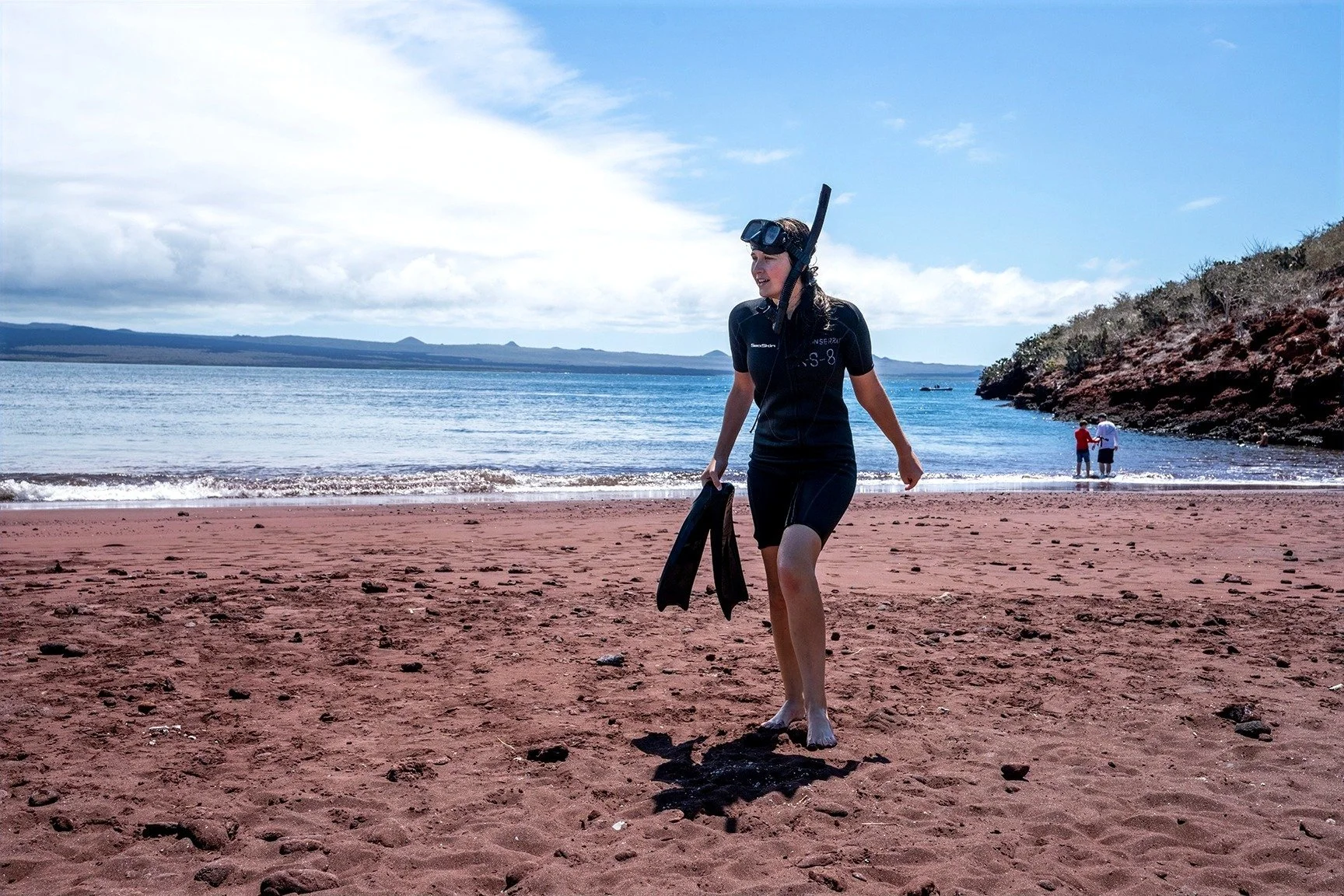 A woman in a wetsuit and snorkel gear walking on a sandy beach carrying flippers, with a view of the ocean, rocks, and two other people in the background.
