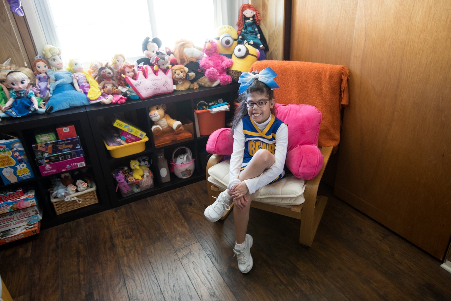 A young girl sitting on a chair, smiling, wearing a cheerleading uniform with a large blue bow in her hair, surrounded by dozens of plush toys on a shelf and a window in the background.