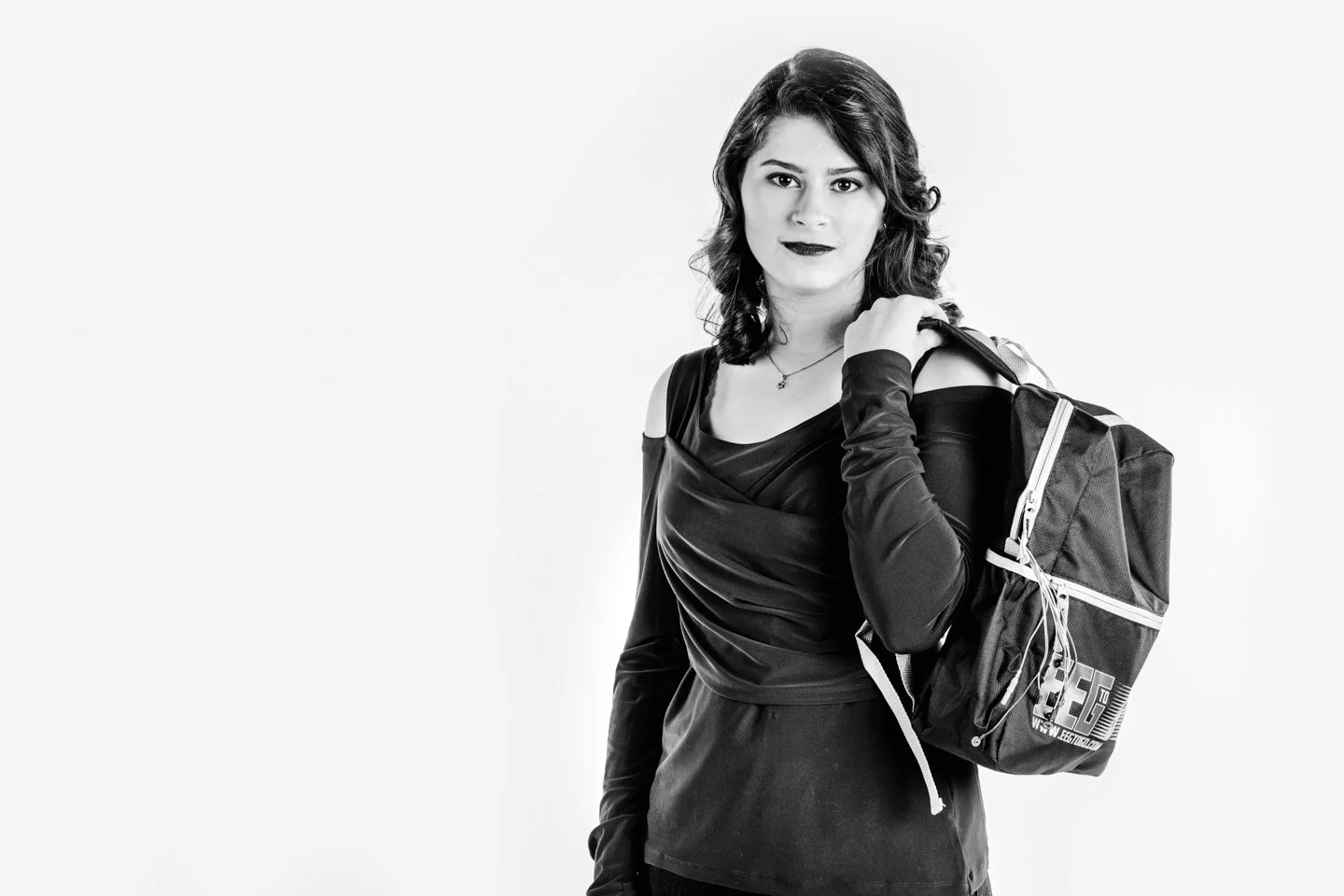 A young woman with wavy hair holding a backpack over her shoulder, wearing a stylish off-shoulder top and long gloves, standing against a plain background.
