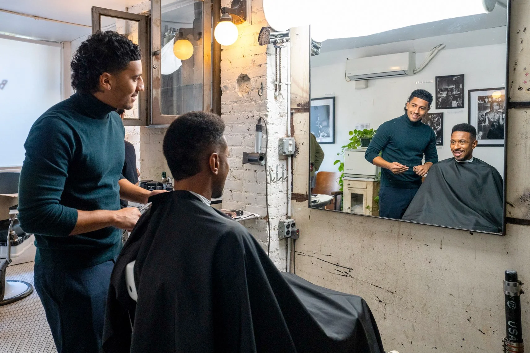 A barber cuts a man's hair while another barber looks on and smiles, as they all interact in a modern barbershop with a mirror, framed pictures, and plants.