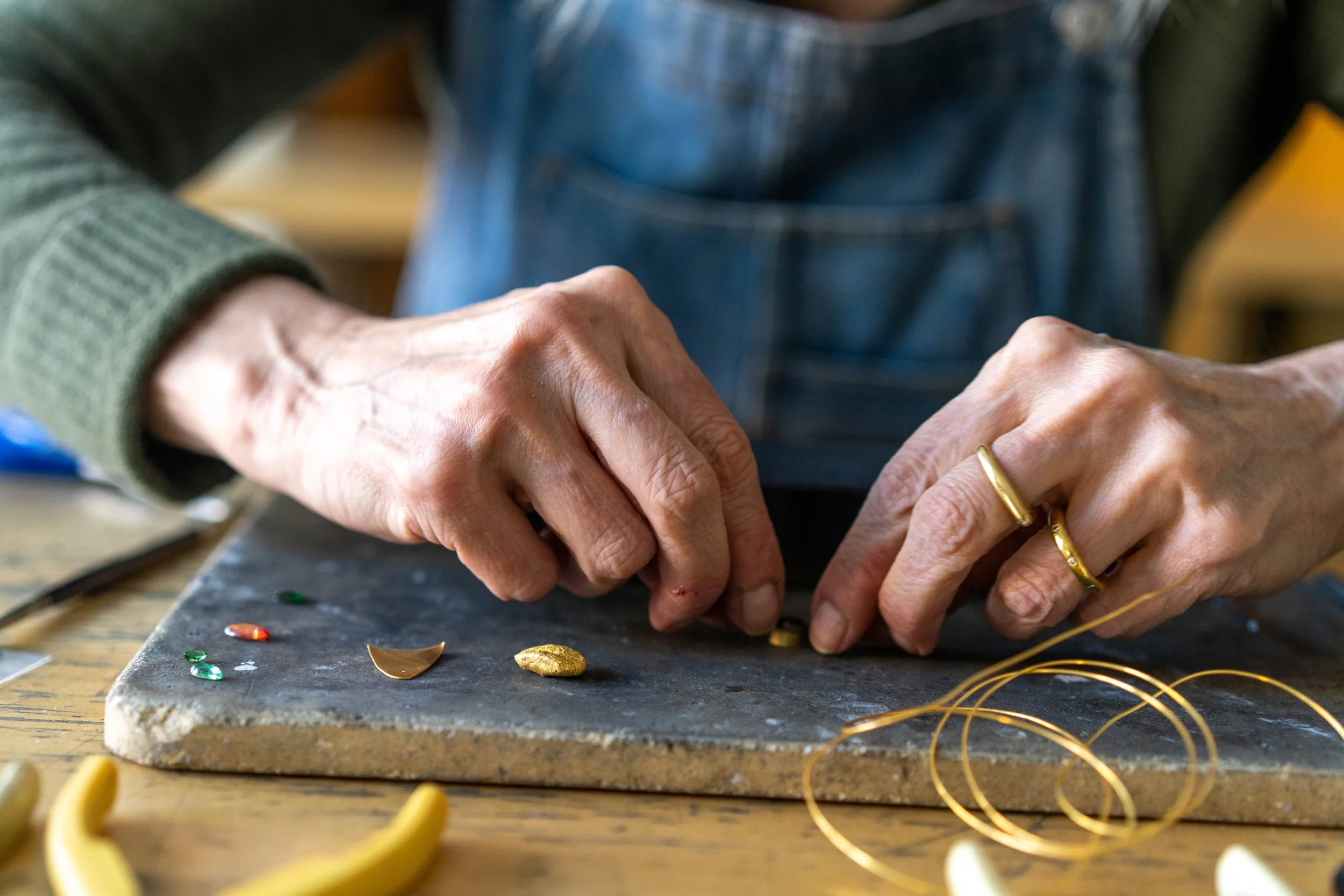 Close-up of a person working on jewelry, using tools to set stones on a dark surface, with gold rings and jewelry items nearby.