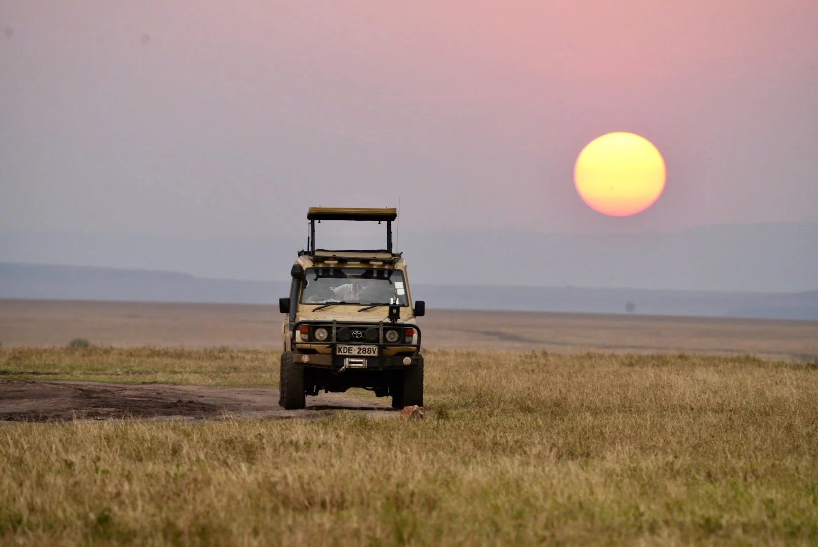 A safari vehicle on a dirt path in an open grassland, with the sun setting in the background and a pinkish sky.