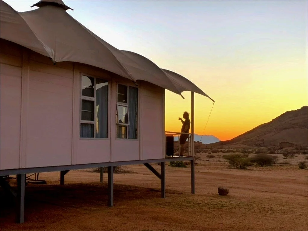 A person standing on a small balcony of a pink house with large umbrellas, during sunset in a desert landscape with mountains in the distance.