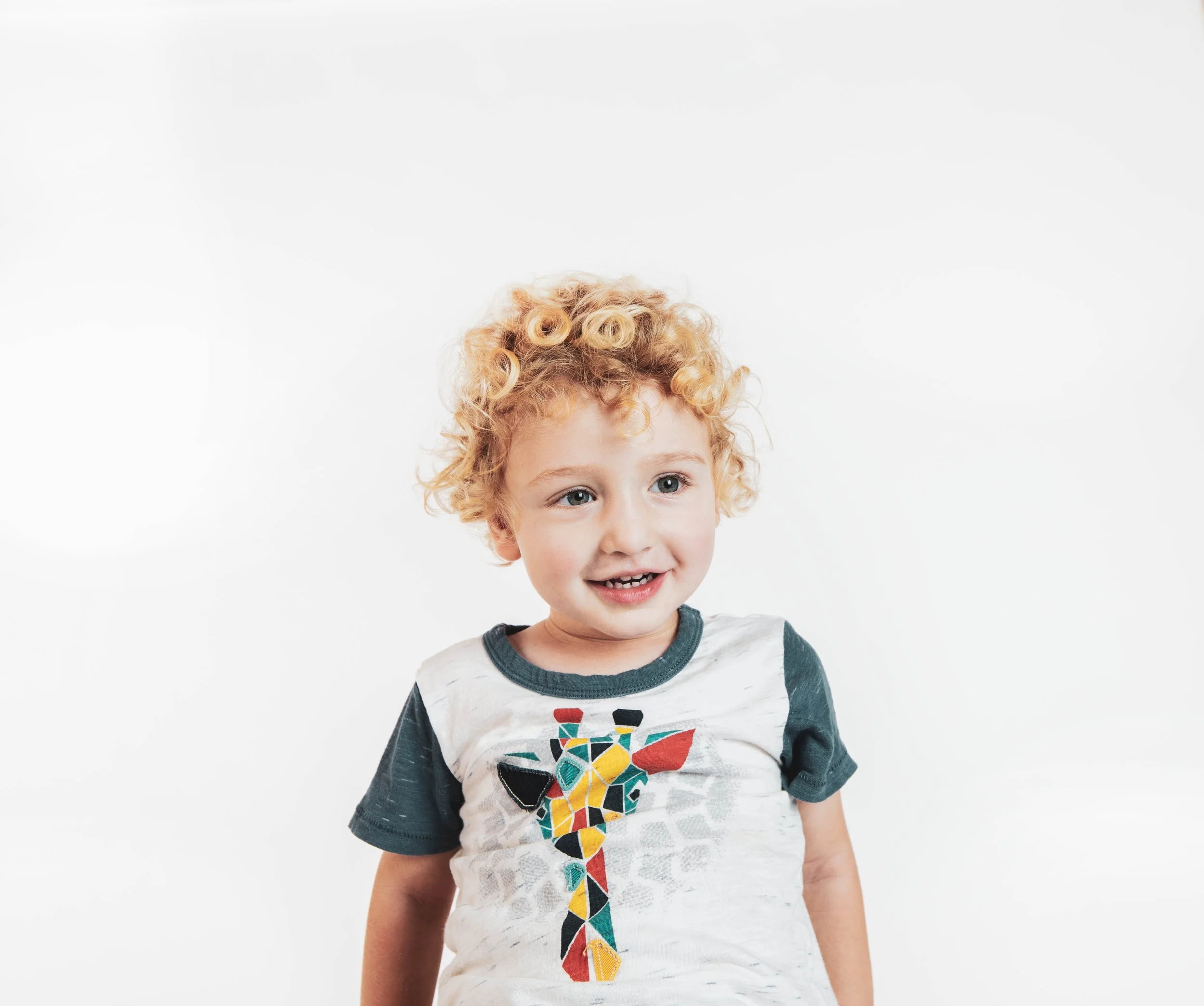 A young boy with curly blond hair and blue eyes smiling, wearing a white and gray T-shirt with a colorful giraffe graphic on the front, standing against a plain white background.