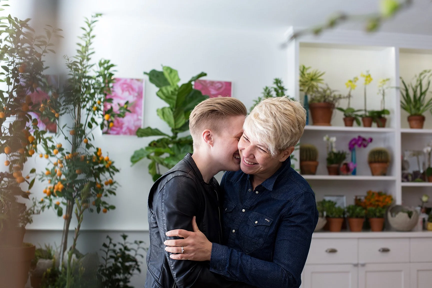 A young woman and an older woman are hugging and smiling at each other in a room filled with potted plants and flowers.