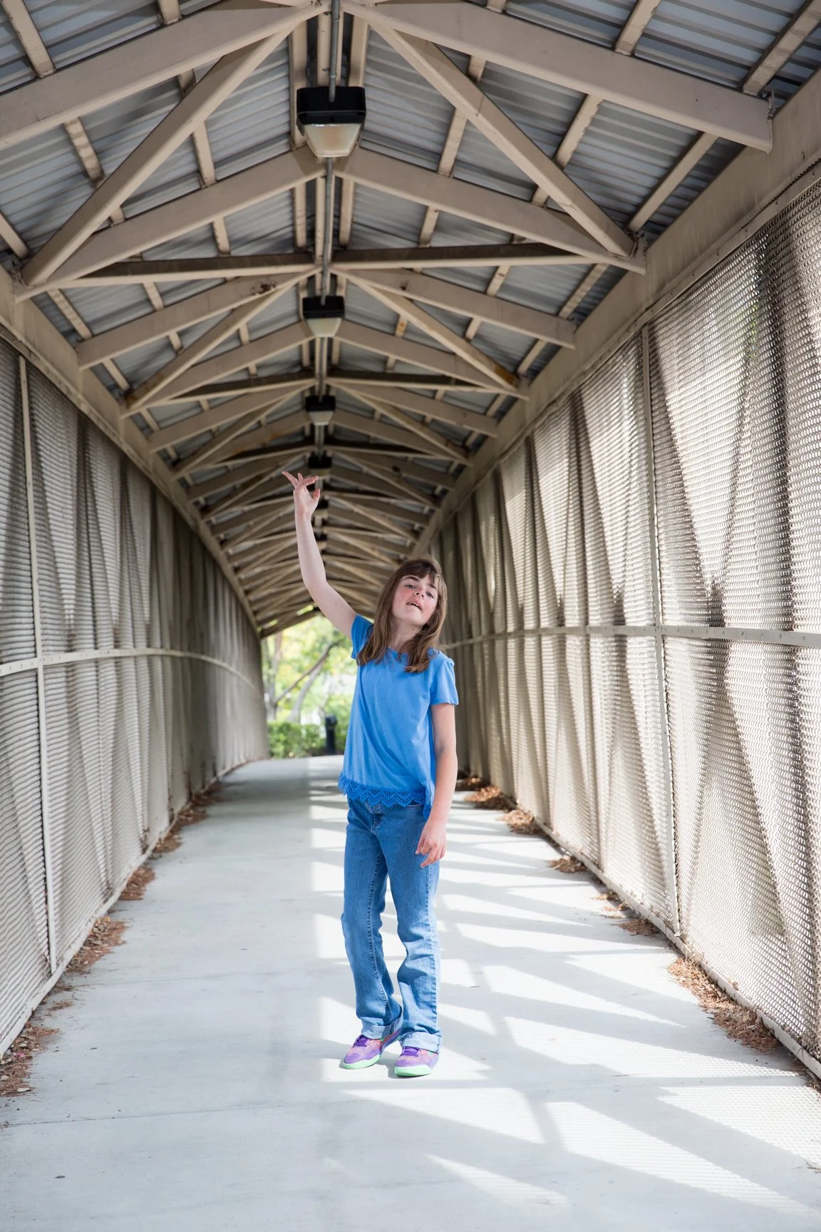 A young girl in a blue shirt and jeans standing in a covered walkway, reaching up with her right hand.
