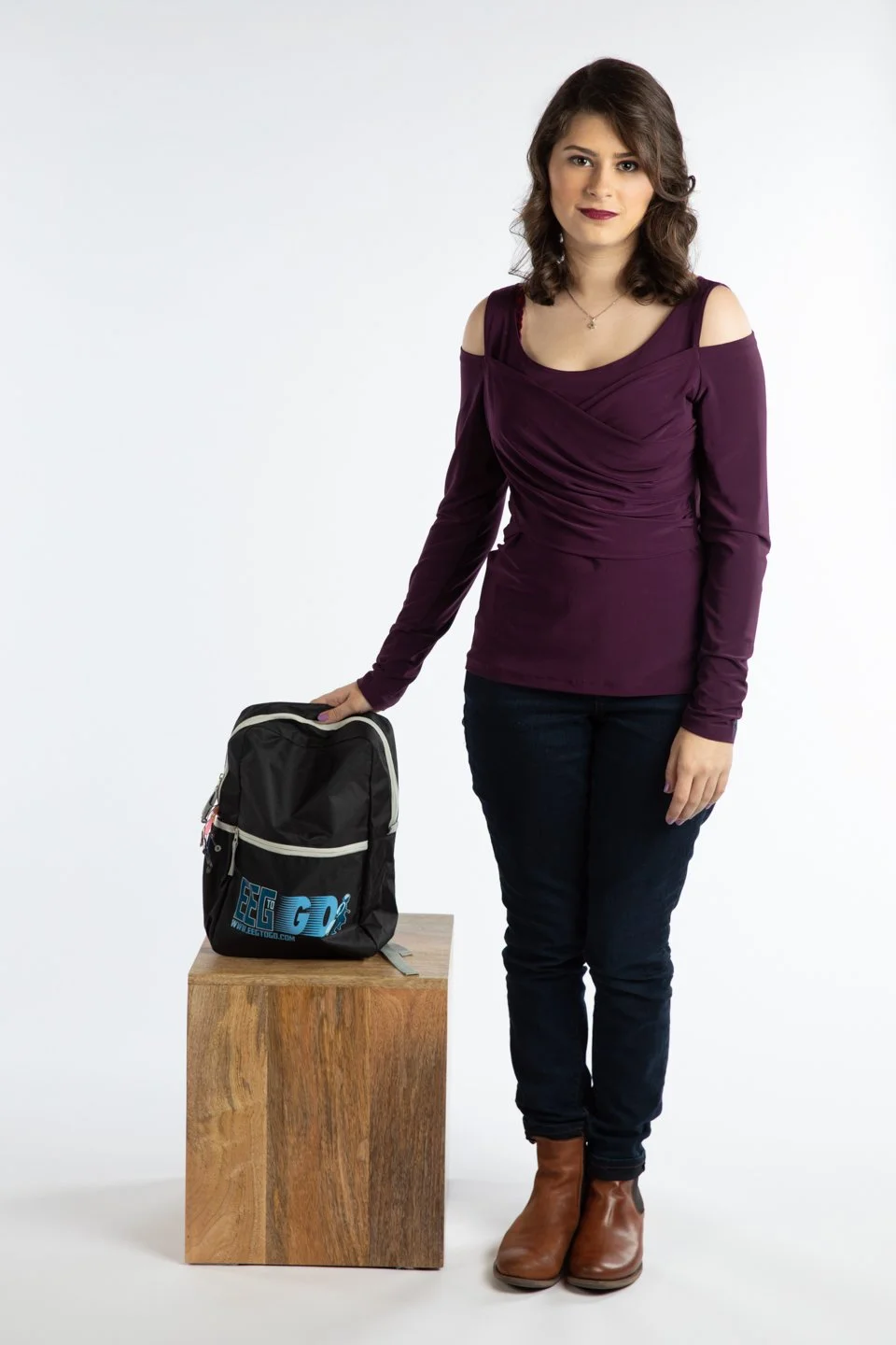 A young woman with brown hair in a purple cold-shoulder top and dark jeans standing next to a wooden block with a black backpack on top, against a white background.