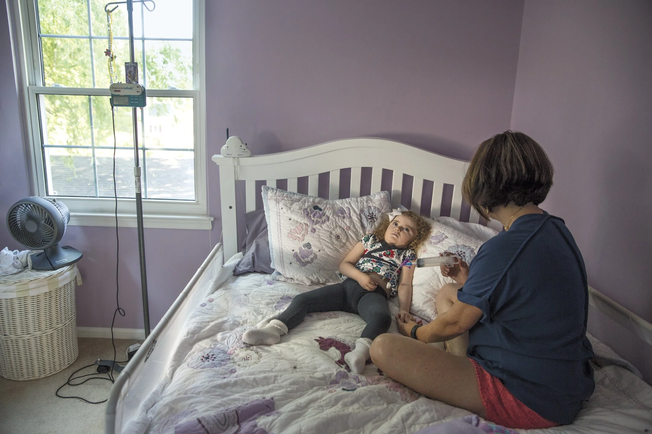 A young girl lying in bed in a purple-walled room, with a woman sitting beside her holding a syringe. The girl looks up at the woman. There is a window with a view outside, a fan on a basket, and an IV pole nearby.