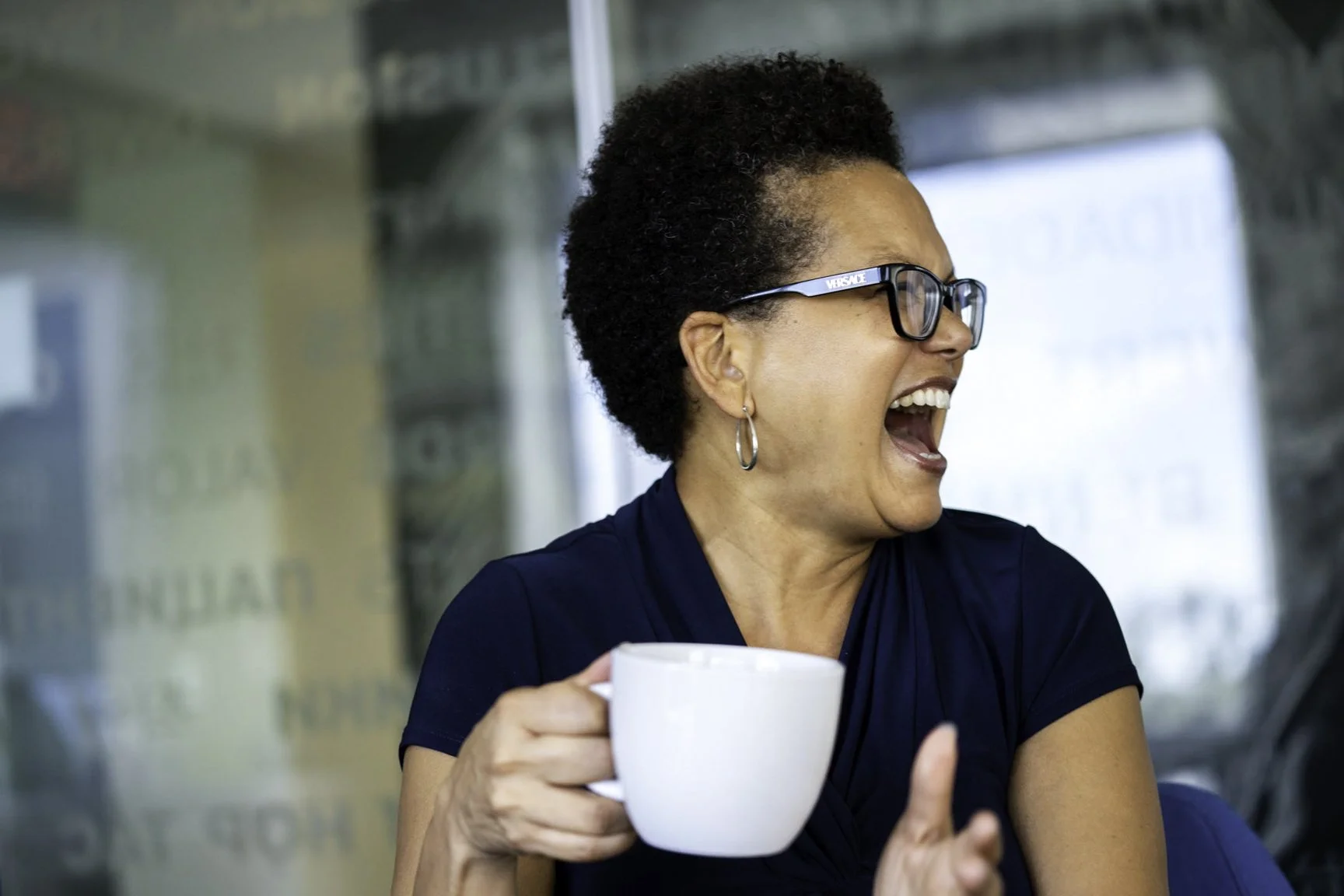 Woman with short curly hair and glasses, smiling and holding a white mug, sitting in a bright indoor space.