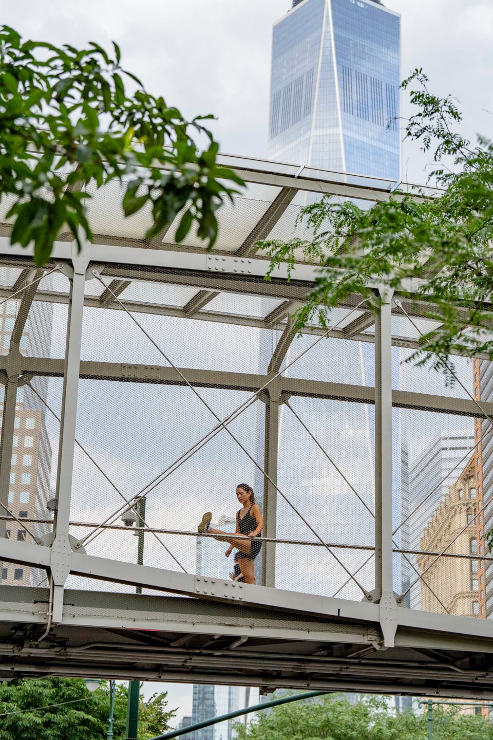 A woman stretching on a pedestrian bridge with a cityscape and tall skyscraper in the background, surrounded by green trees.