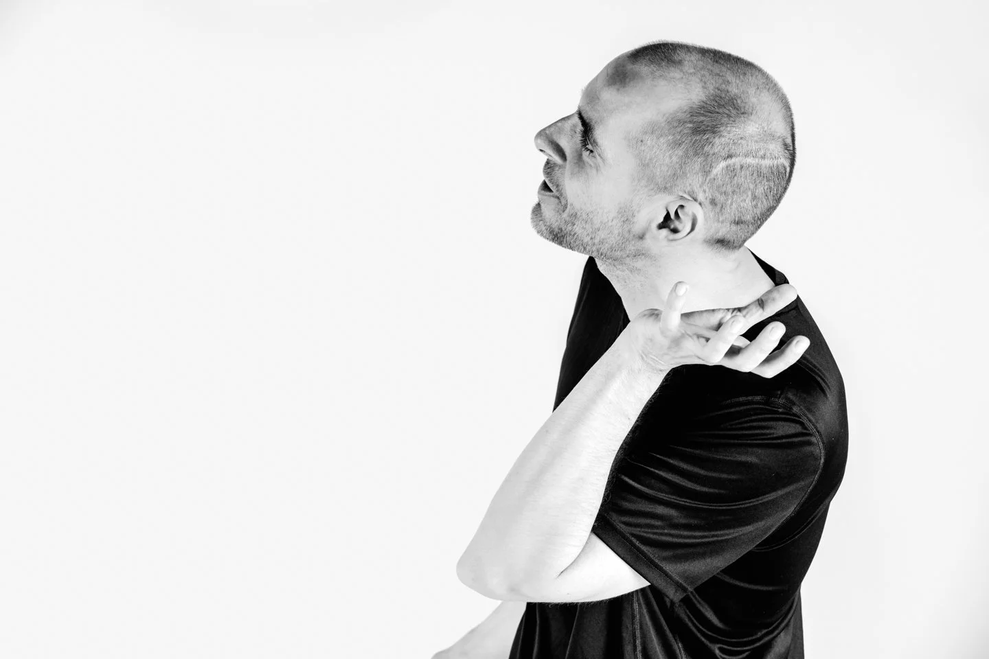 Black and white photo of a man with a shaved head and beard, wearing a black t-shirt, lifting his hand near his neck and looking upwards.