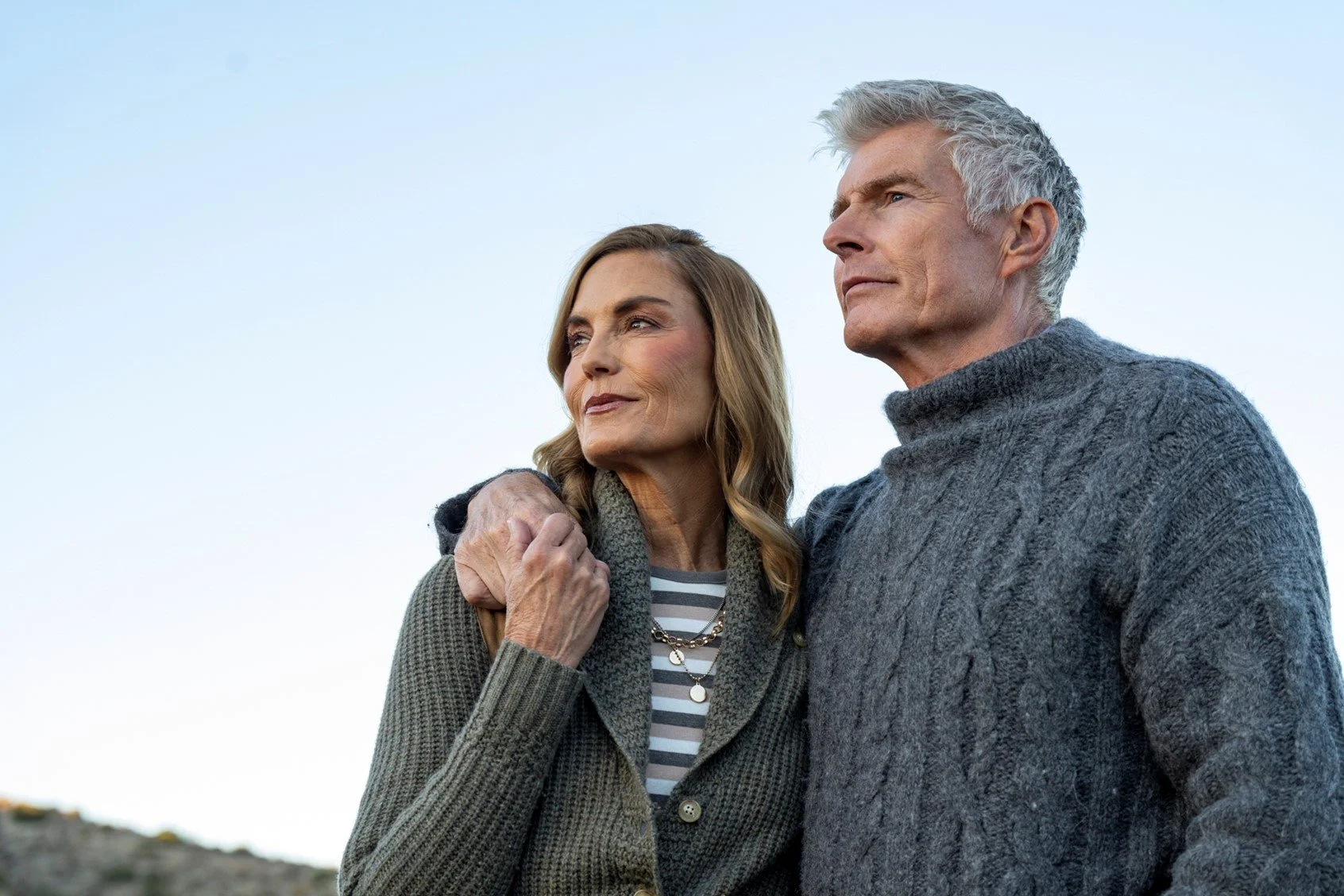 An older man and woman stand close together outdoors, with the man’s arm around the woman’s shoulder. Both are dressed in warm sweaters and gazing into the distance with serious expressions.