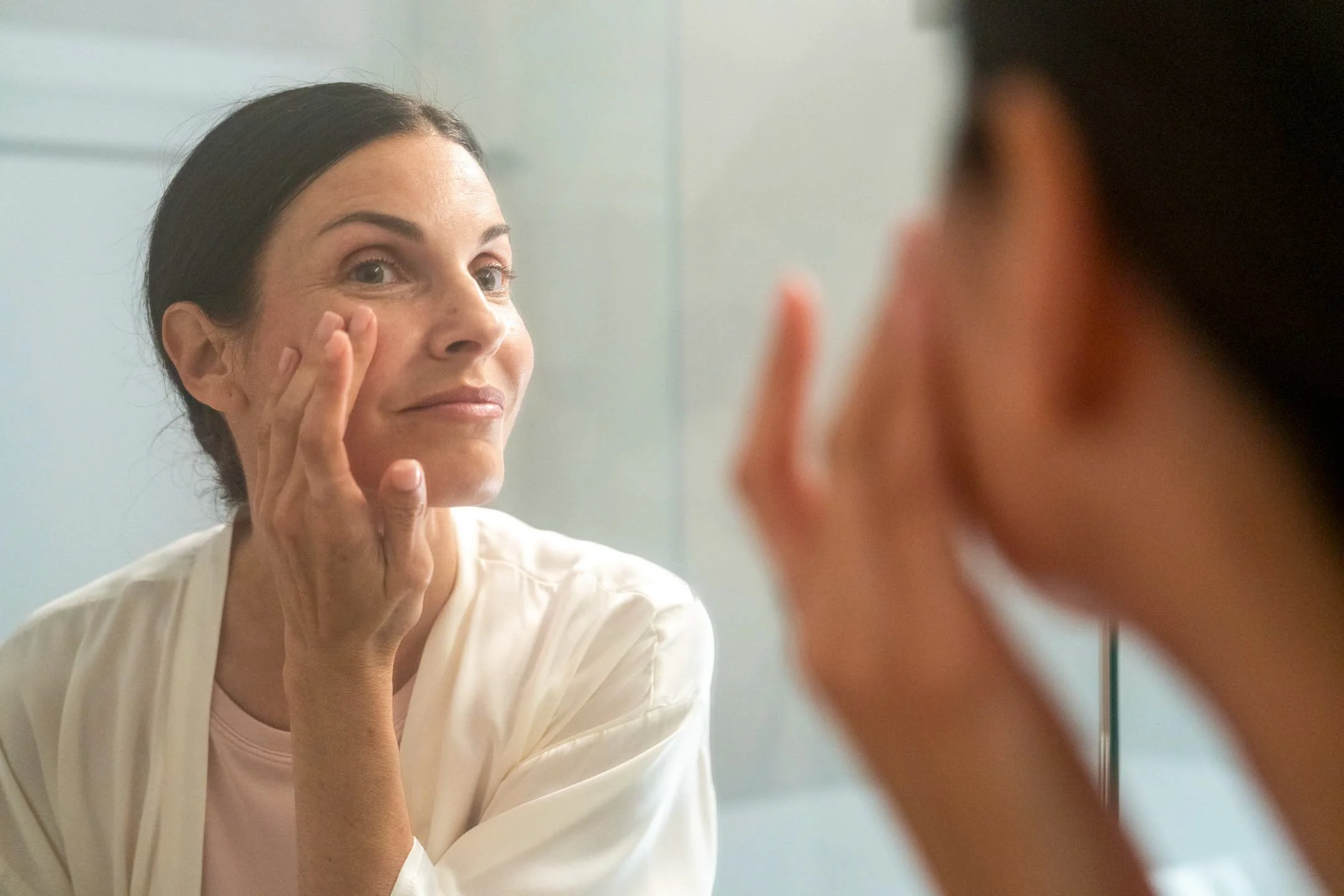 A woman with dark hair looking at her reflection in a mirror while touching her face.