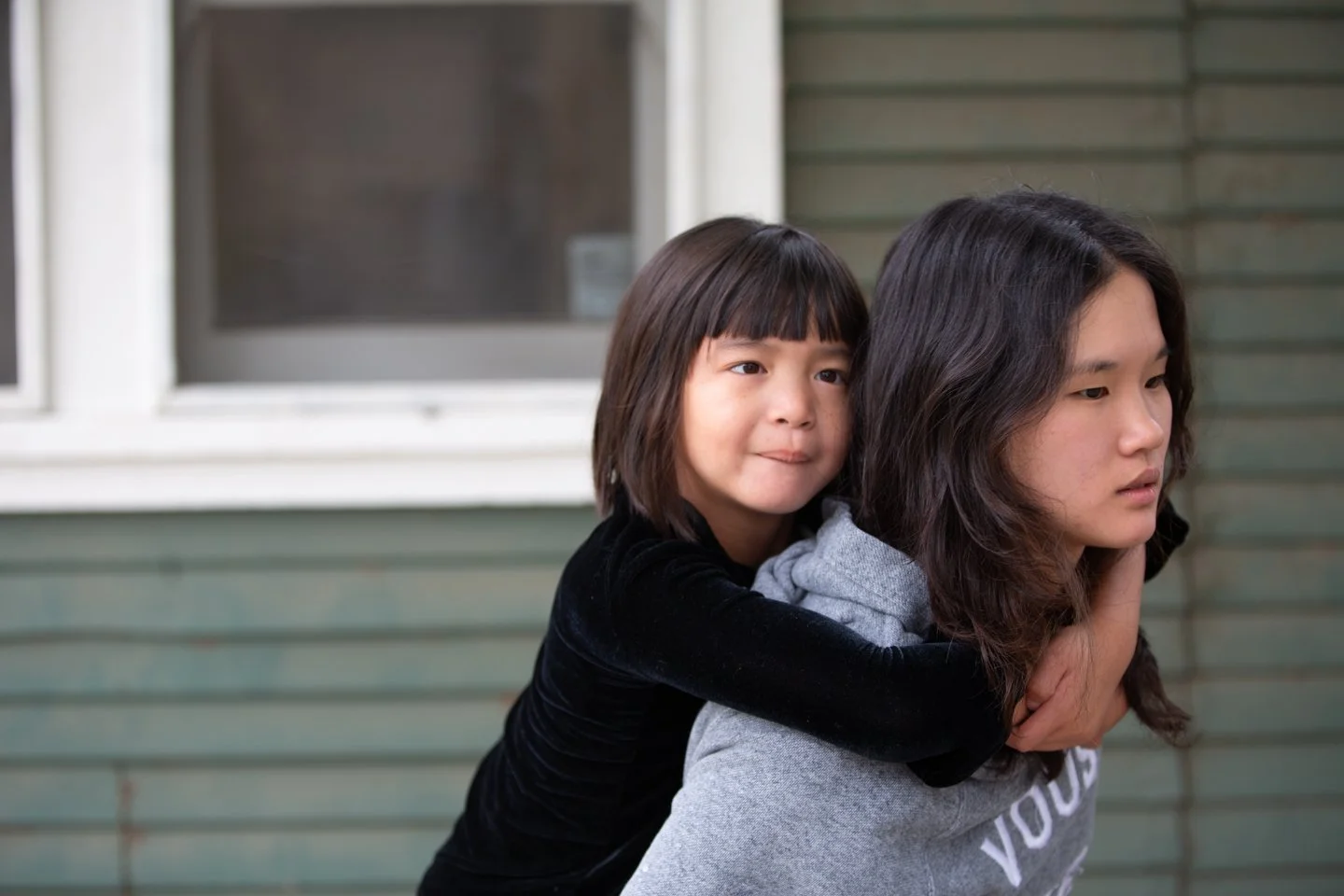A young girl with short brown hair giving a piggyback ride on a woman with long dark hair, no glasses, and a grey hoodie, in front of a house with green siding and a window.