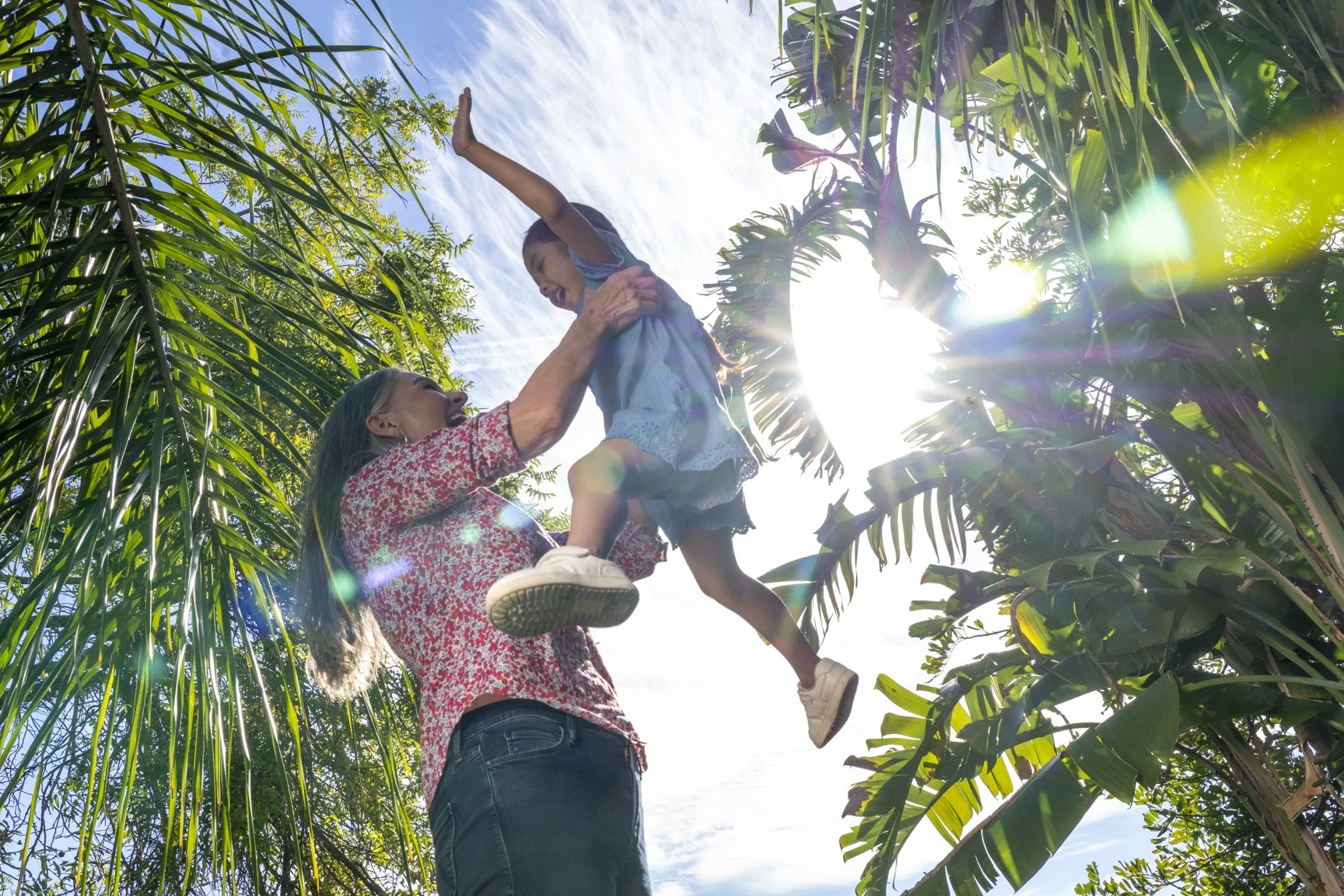 A woman lifting a young girl in a tropical orchard with green palm trees and banana plants, and bright sunlight in the background.