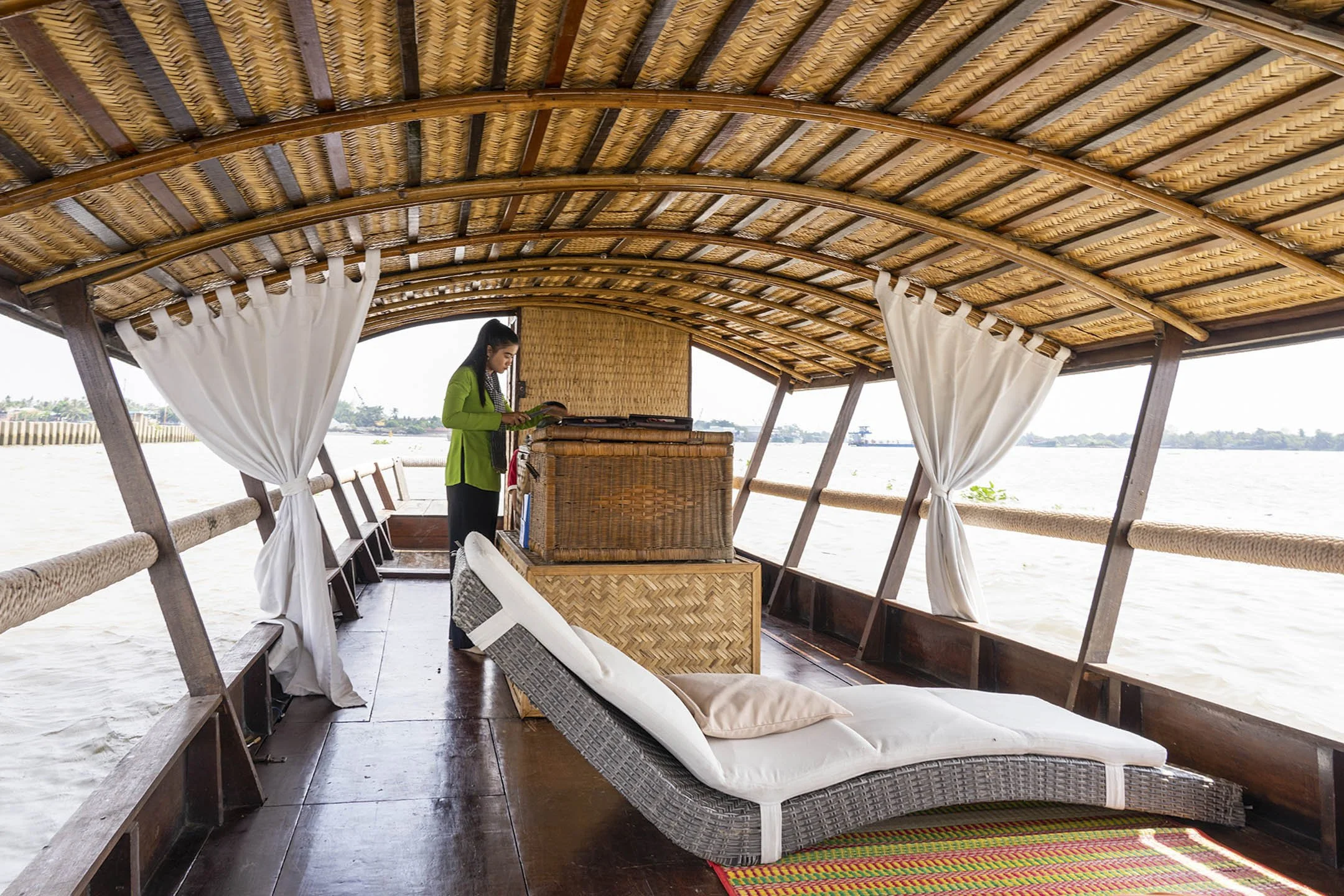 Woman on houseboat with wicker furniture and white curtains by water.