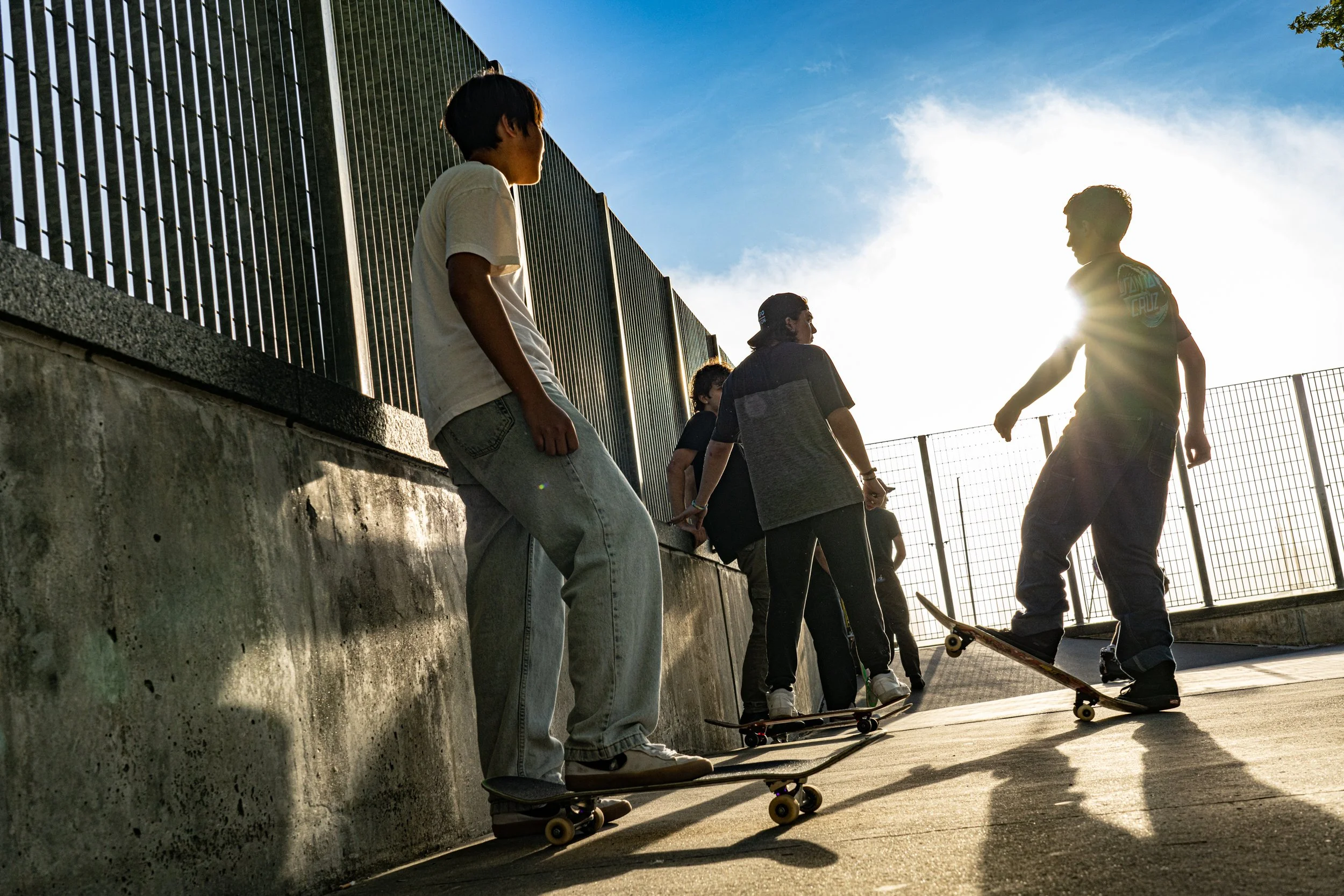 A group of young skateboarders hanging out by a concrete ledge at sunset, with a metal fence and clear sky in the background.