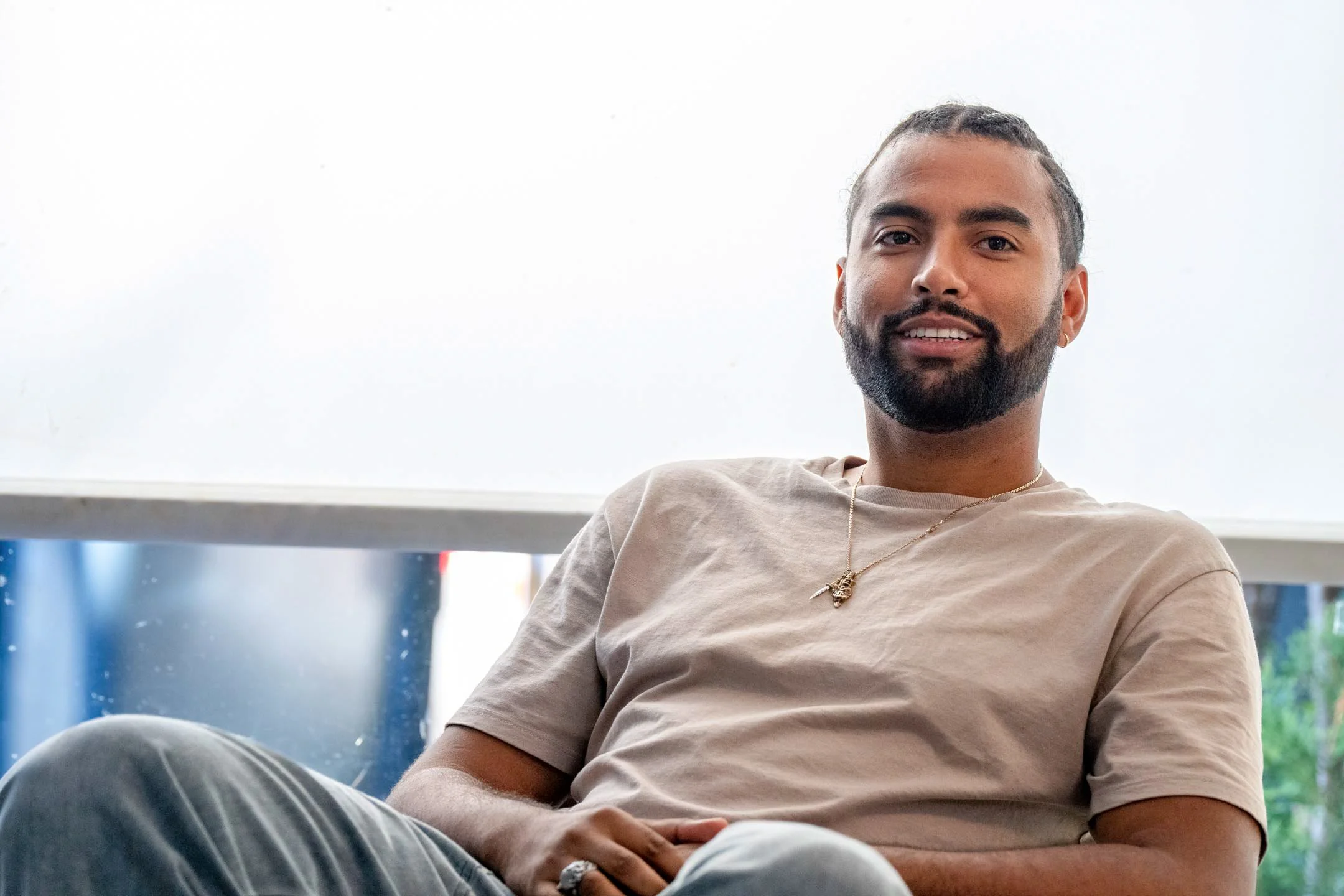 A young man with dark hair, a beard, and a small earring, sitting comfortably and smiling in front of a large window with a blurry outdoor view.